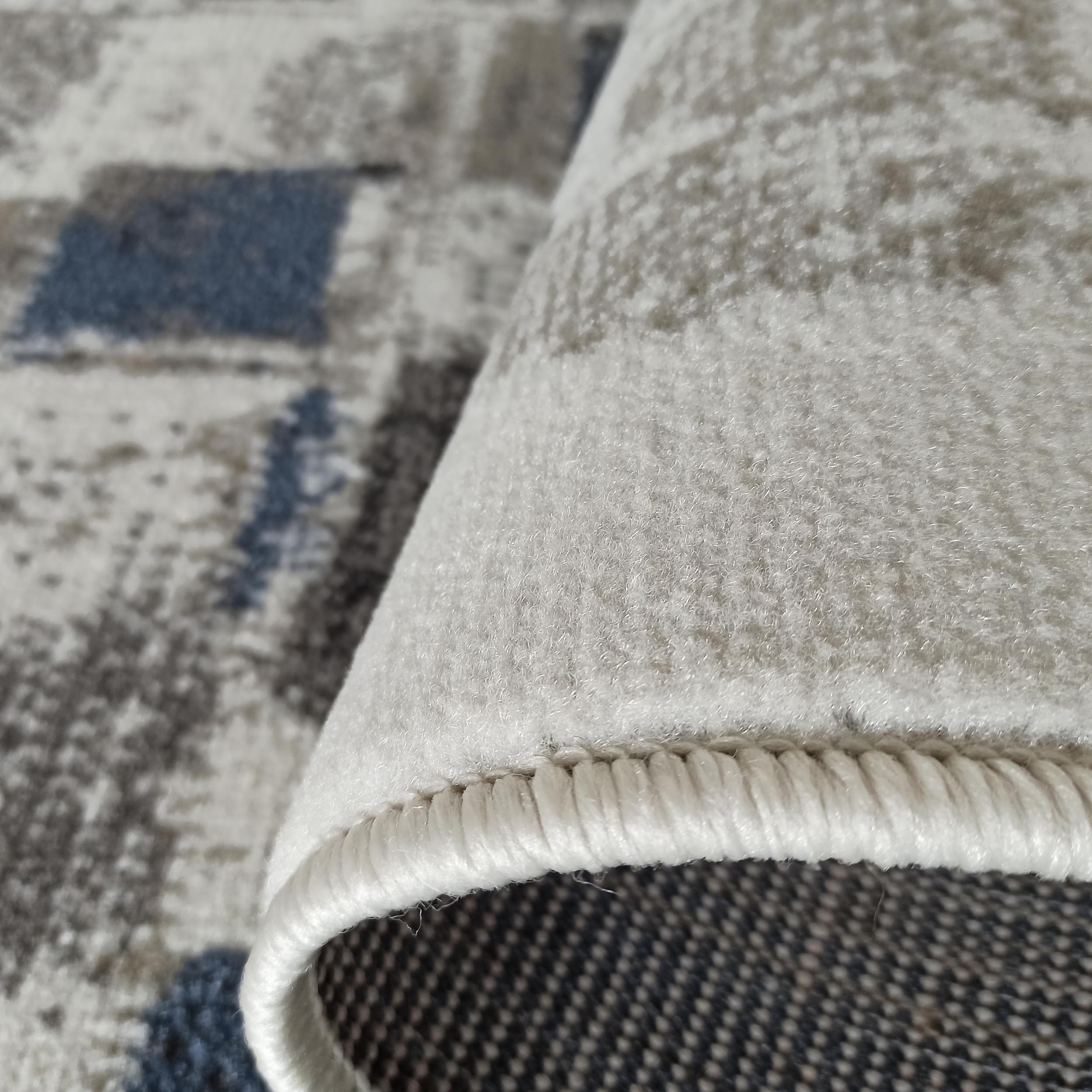 Gros plan sur un tapis à poils doux, avec des motifs abstraits en gris, bleu et blanc cassé. Le tapis est partiellement roulé, révélant son envers tissé foncé