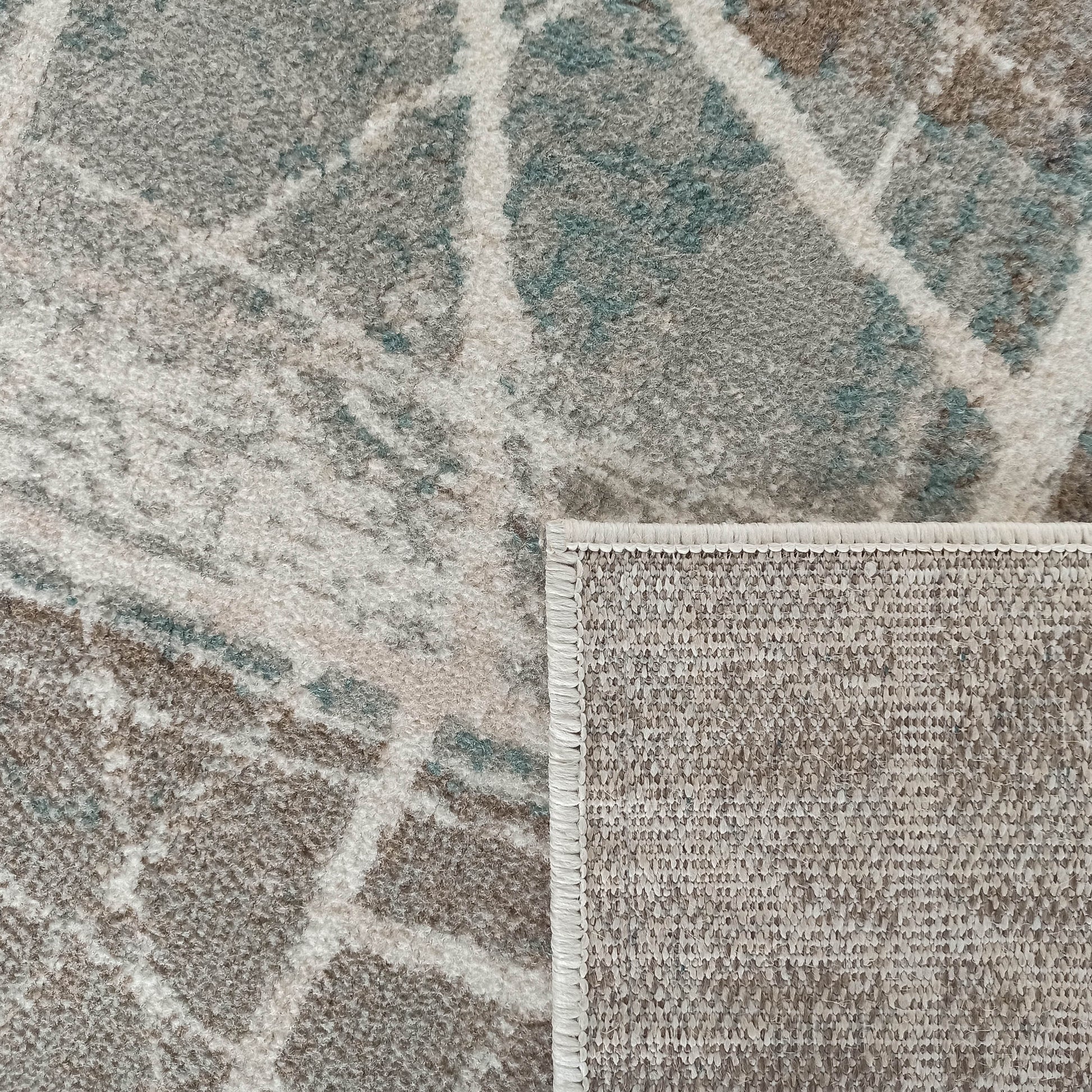 Tapis texturé aux motifs abstraits gris, beiges et bleu-vert, avec un coin relevé montrant l'envers tissé