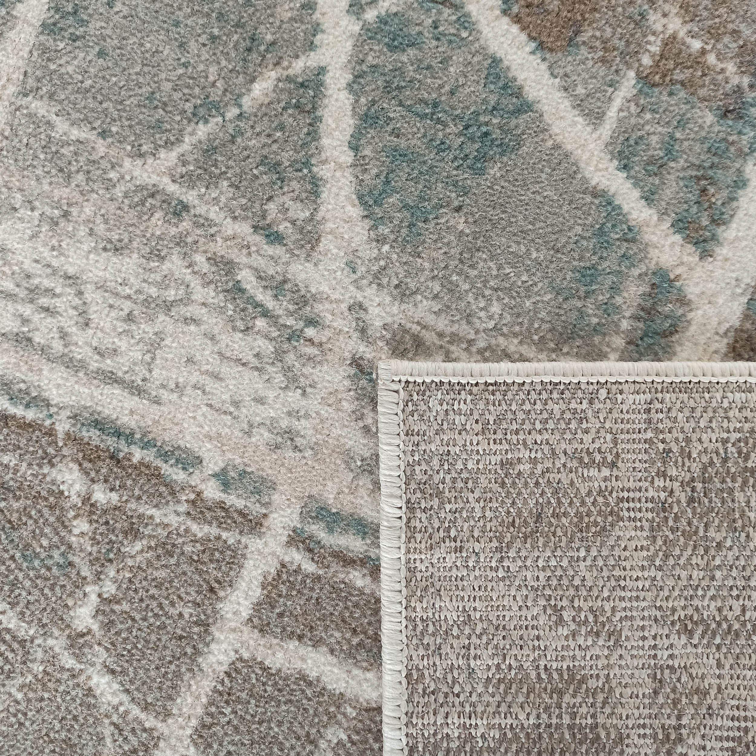 Tapis texturé aux motifs abstraits gris, beiges et bleu-vert, avec un coin relevé montrant l'envers tissé