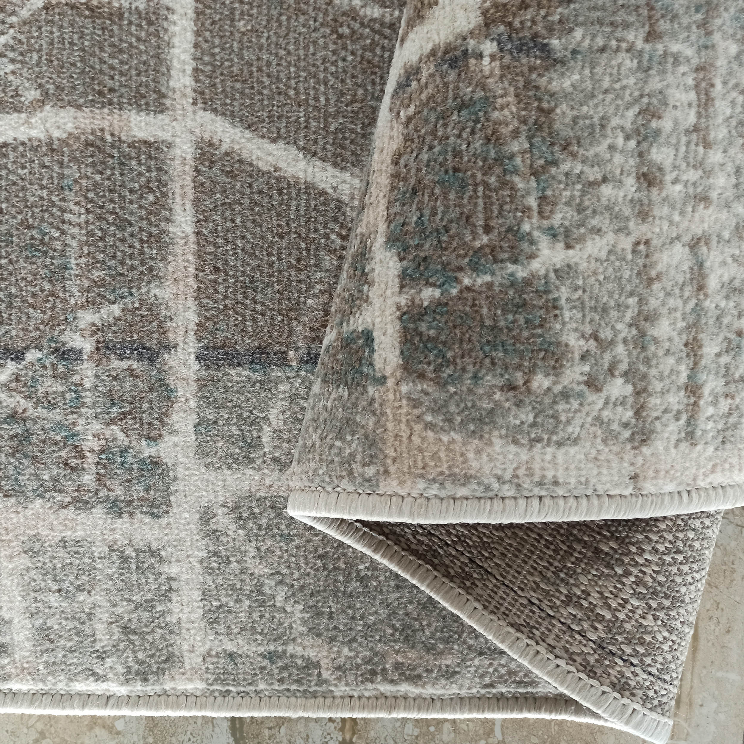 Tapis partiellement roulé aux motifs géométriques abstraits gris, beige, crème et bleu-vert, avec une texture douce