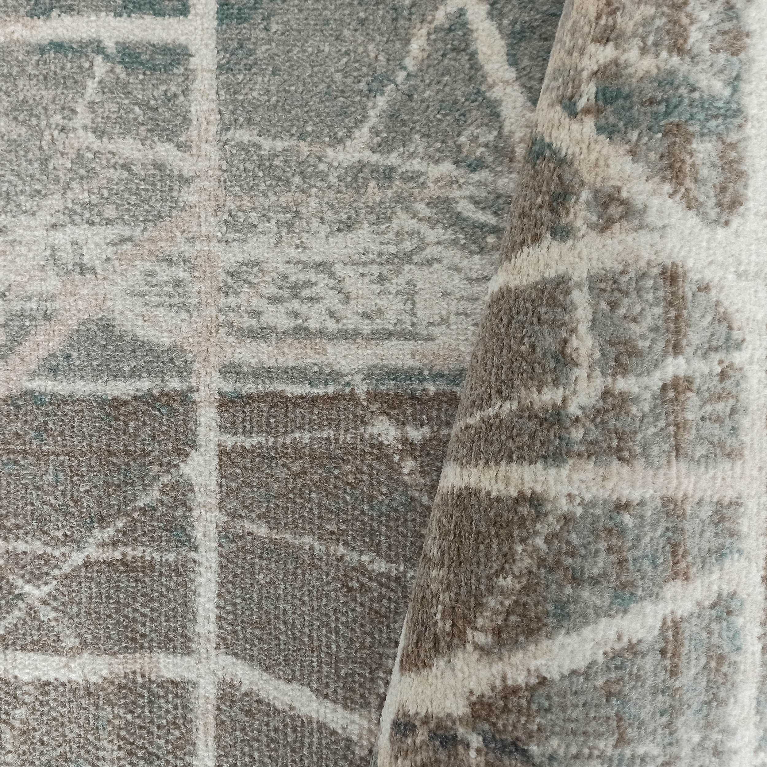 Tapis à texture duveteuse, motifs géométriques abstraits en gris, beige et touches de bleu-vert, en partie roulé