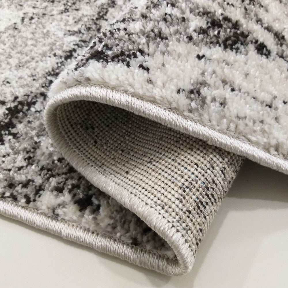 Tapis texturé blanc, gris et noir aux motifs abstraits. Un coin est roulé, révélant l'envers tissé et le liseré gris clair