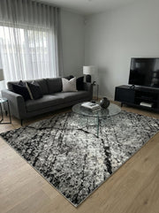 Tapis rectangulaire à motifs abstraits noirs, gris et blancs, créant un effet texturé et patiné sur un sol en bois clair d'un salon