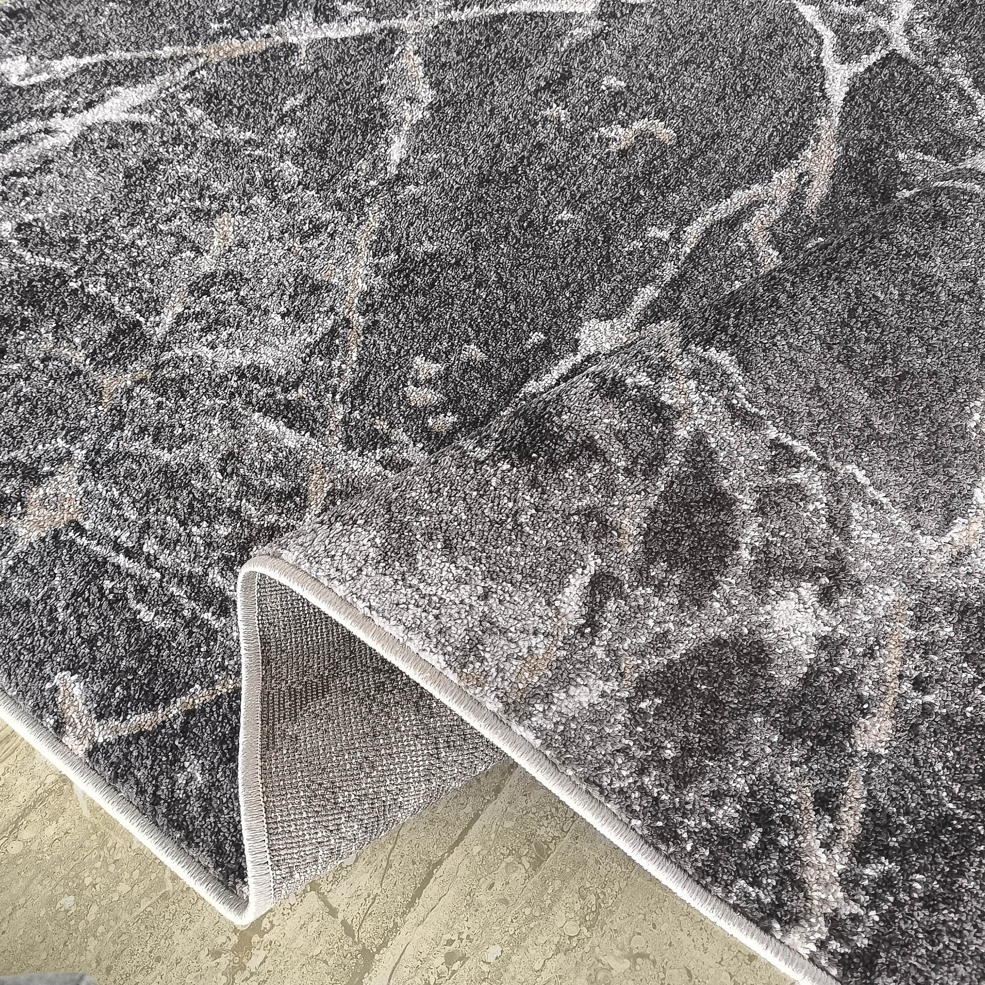 Tapis texturé aux tons gris foncé et clair, marbré de motifs abstraits beiges, replié sur un sol clair révélant sa bordure
