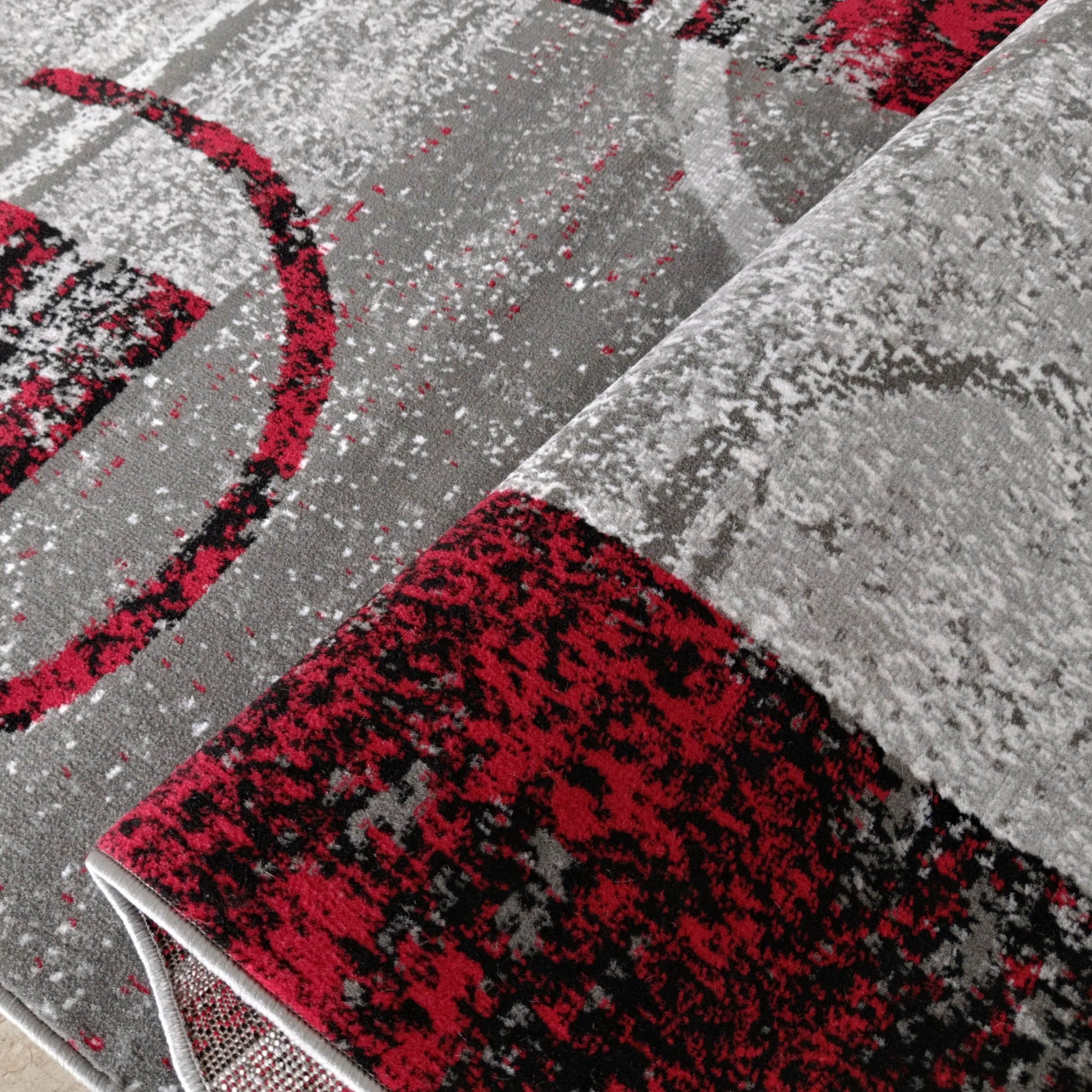 Tapis abstrait moucheté gris, rouge et noir, partiellement roulé révélant un envers tissé et des formes incurvées