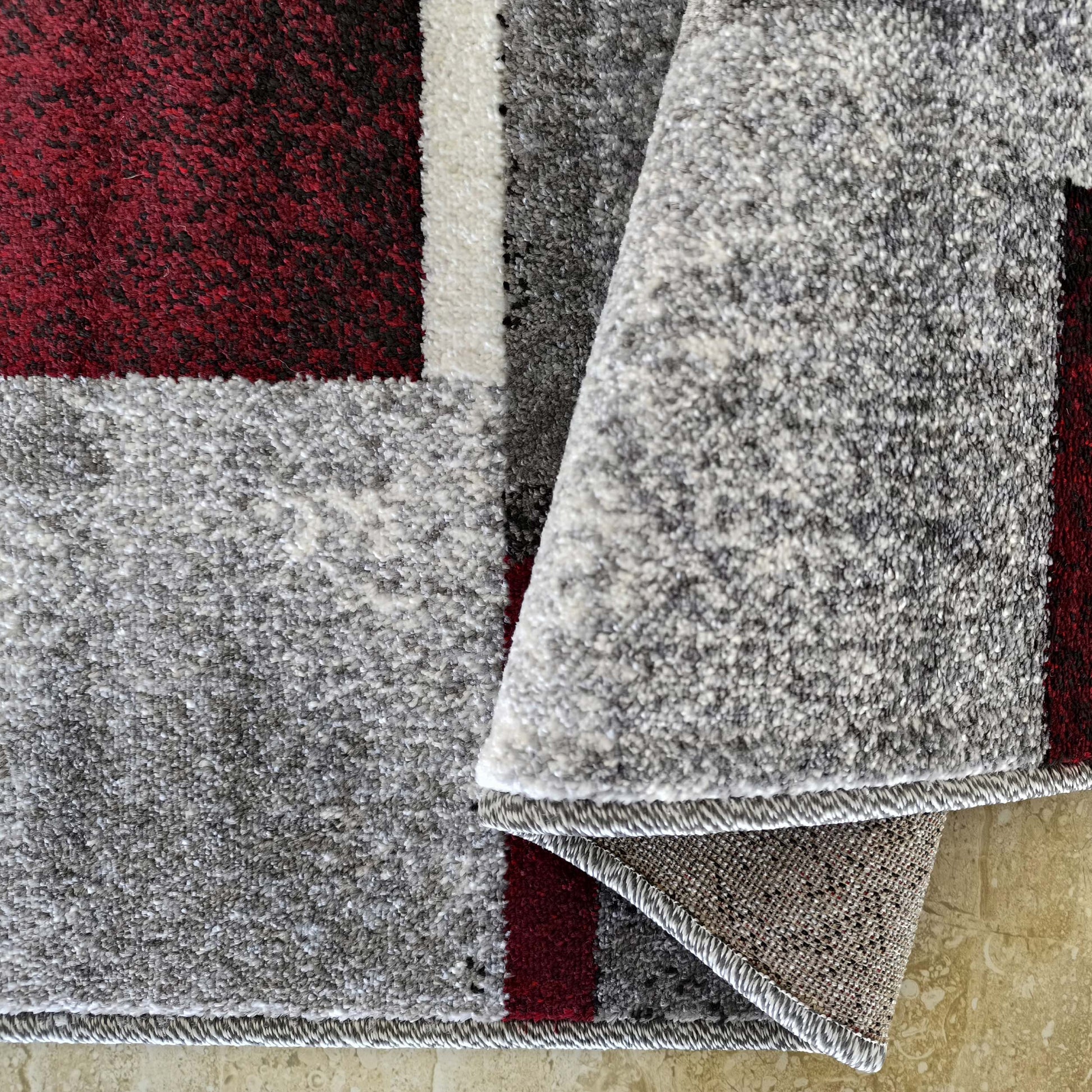 Tapis à motifs géométriques rouge foncé, gris et blanc, avec une texture douce et bouclée, partiellement roulé