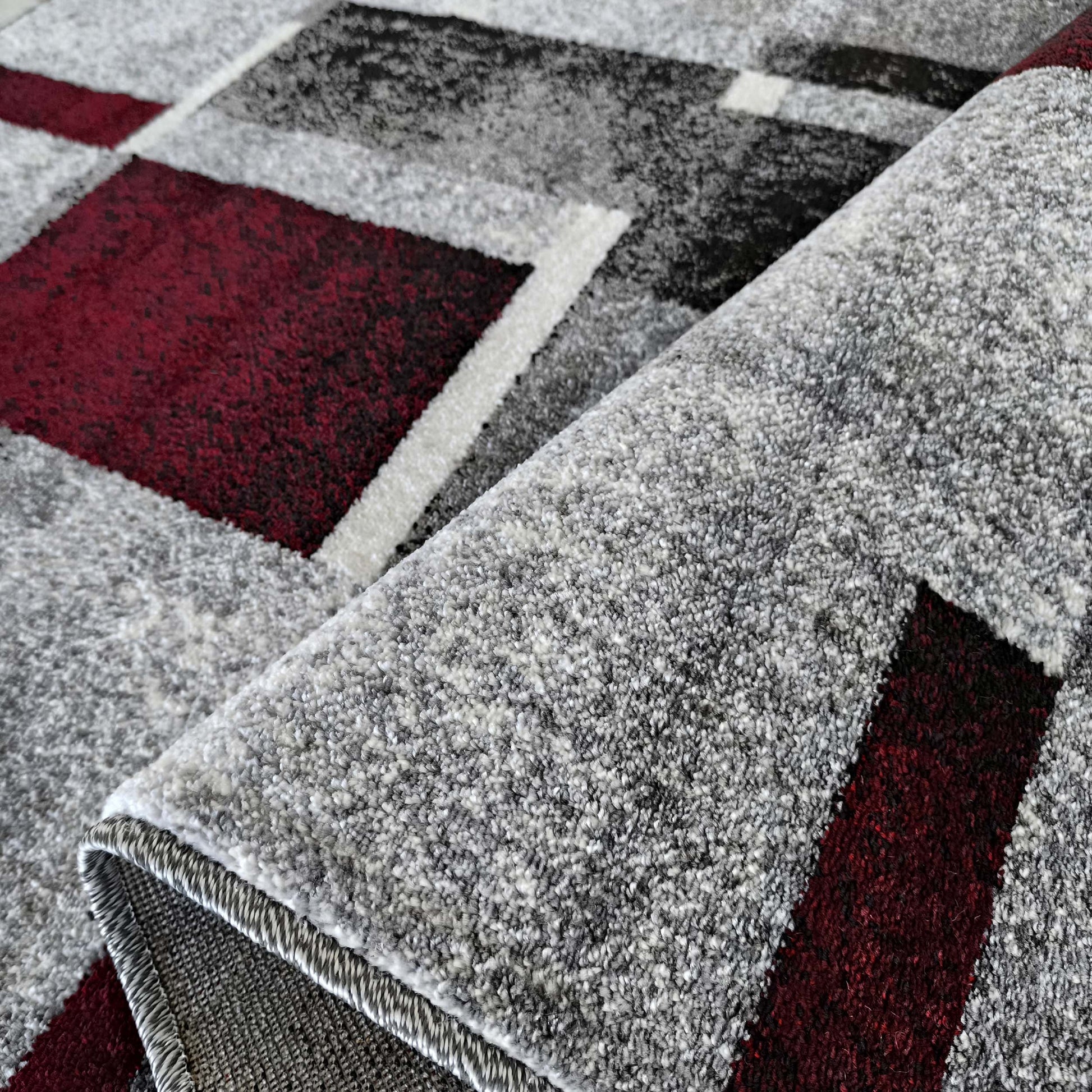 Tapis aux motifs géométriques gris, bordeaux et blanc, présentant une texture chinée et épaisse, dont une partie est roulée
