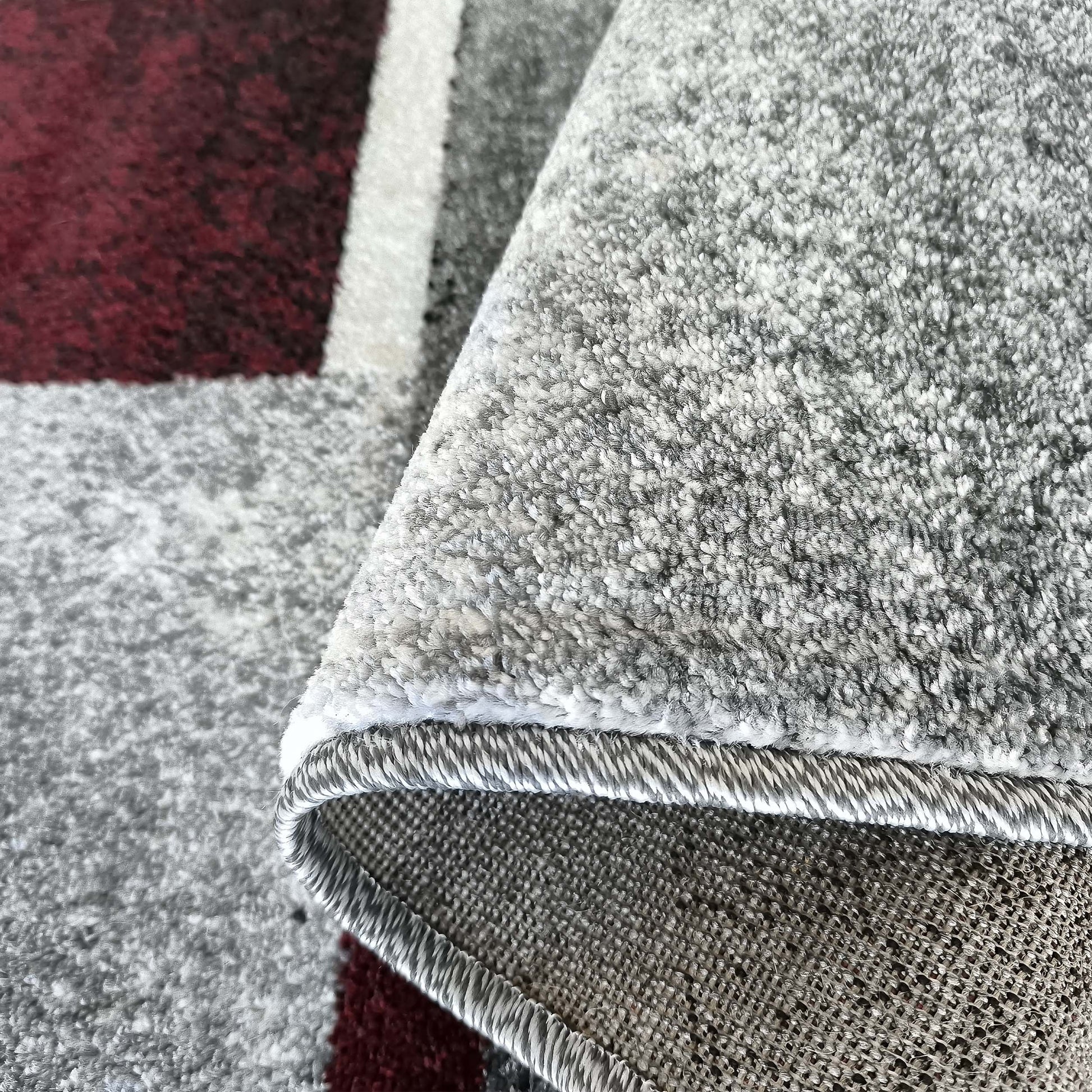 Tapis doux à motifs géométriques rouge bordeaux, gris et blanc, un coin est relevé montrant la texture dense et le dos tissé