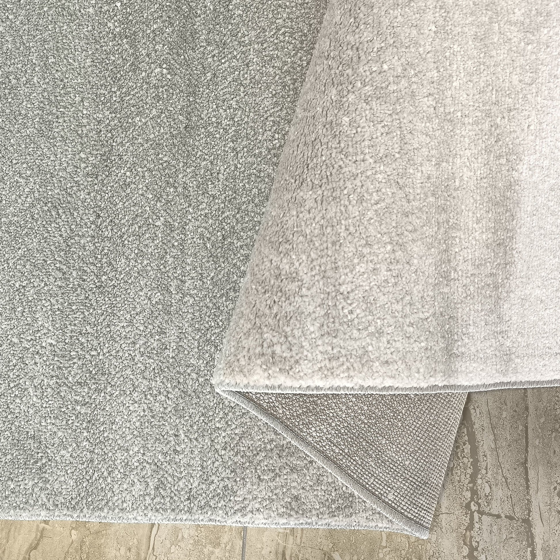 Tapis gris argenté à l'aspect moelleux, un pan est relevé révélant son envers tissé sur un sol beige