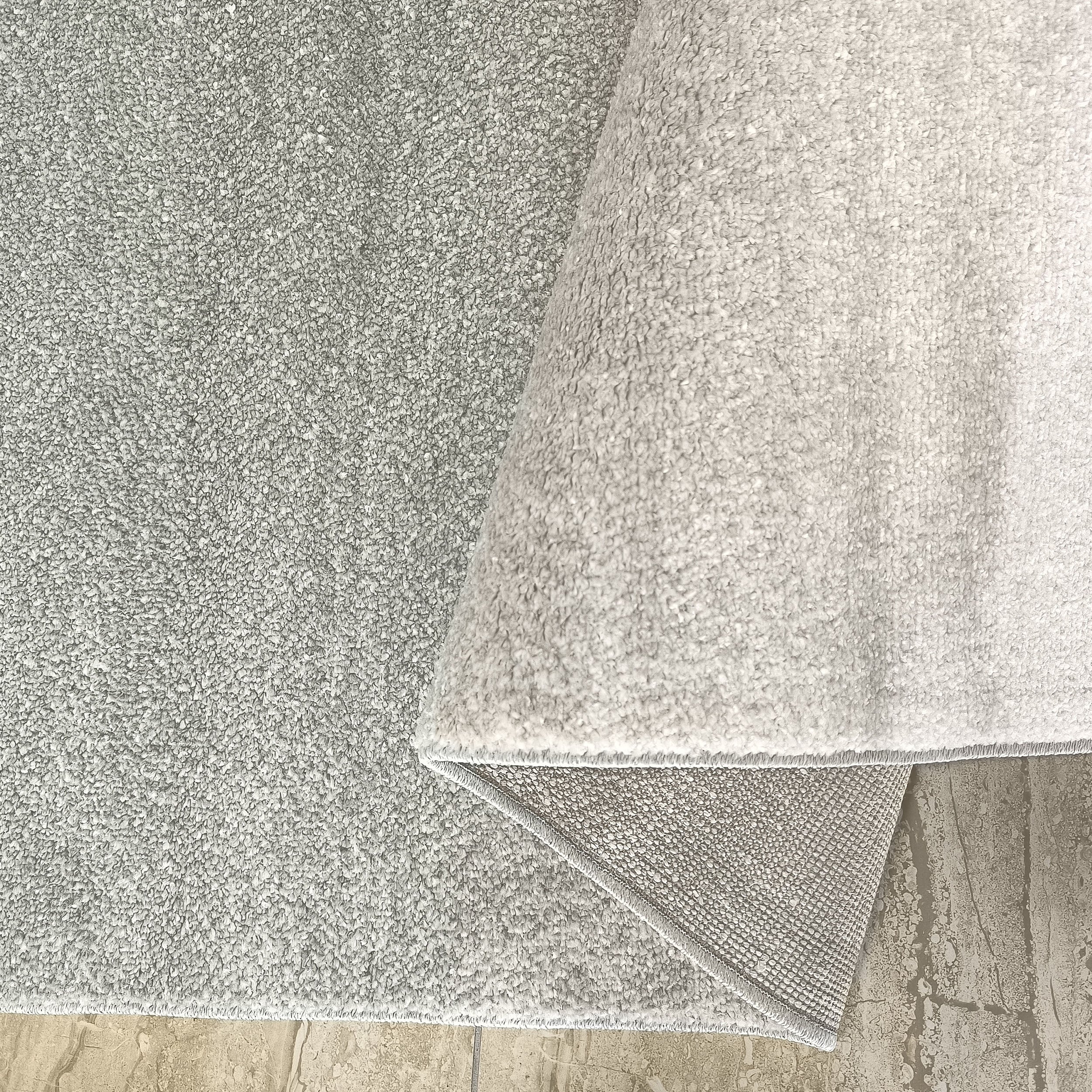 Tapis gris argenté à l'aspect moelleux, un pan est relevé révélant son envers tissé sur un sol beige