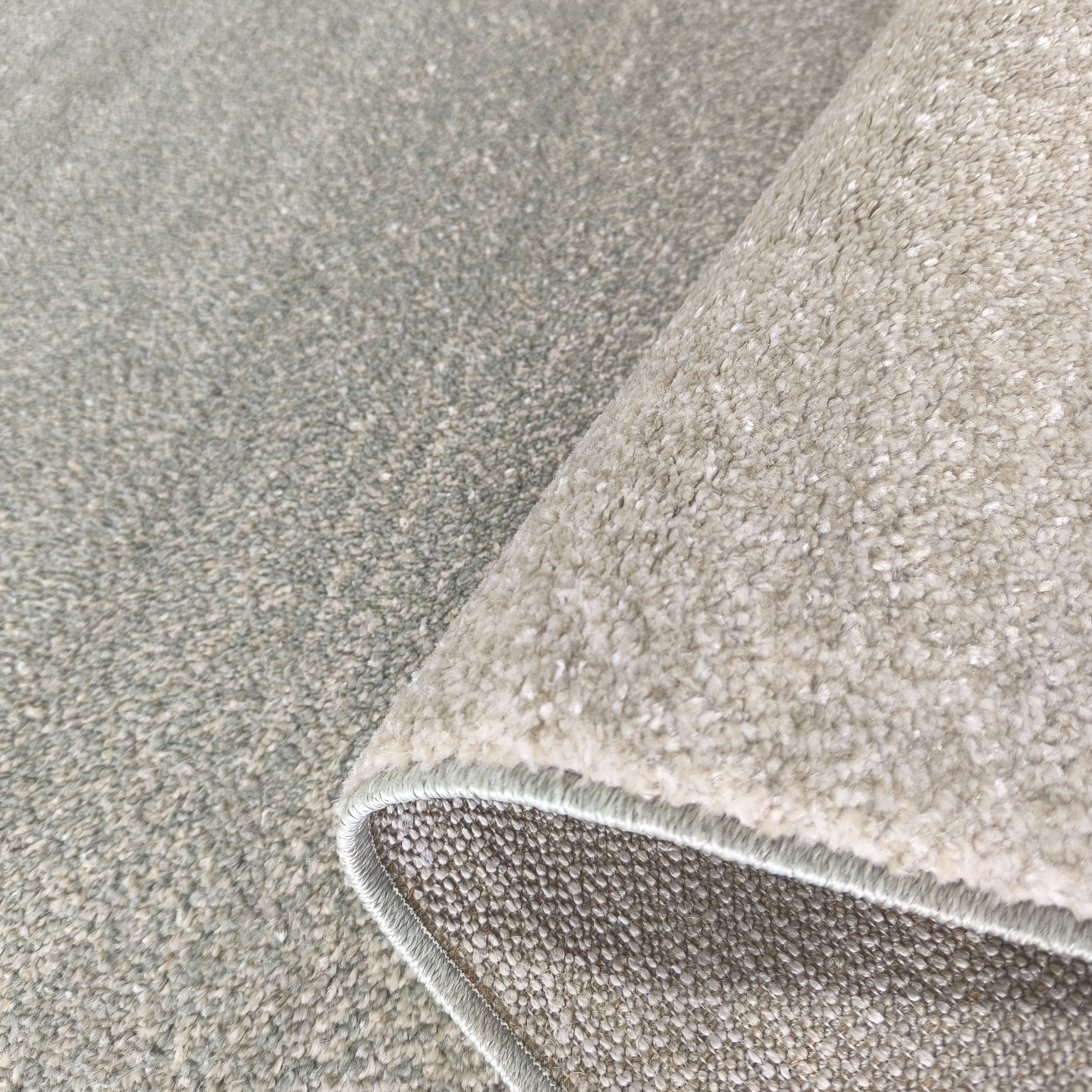 Tapis texturé gris perle et blanc cassé avec un bord roulé, révélant la texture bouclée du dessus et le revers tissé
