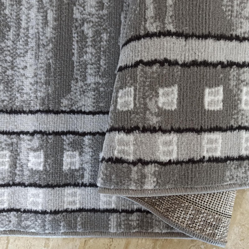 Tapis gris aux motifs abstraits clairs et foncés, avec des rayures noires et des carrés, partiellement plié