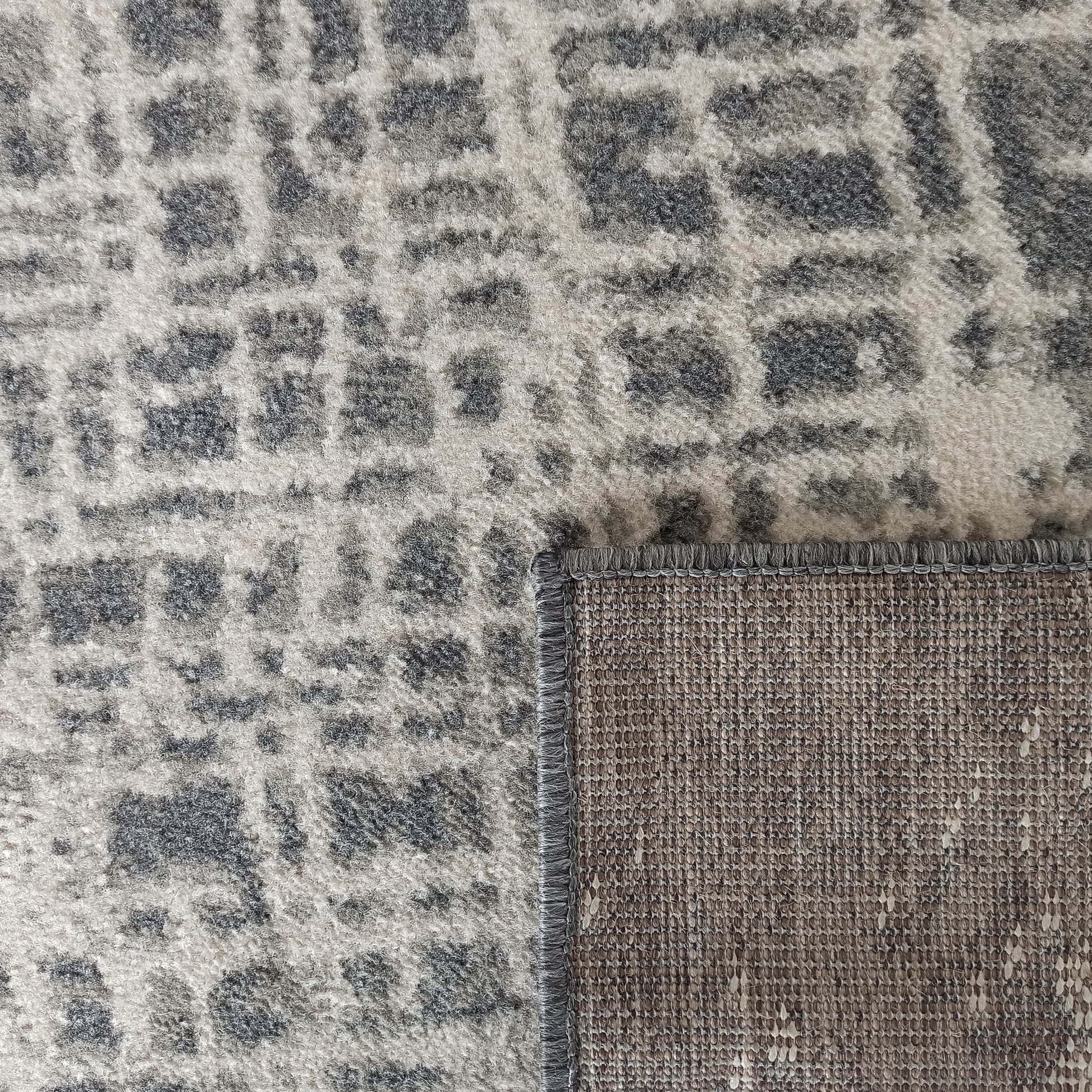 Tapis à motif abstrait gris clair et foncé avec une surface moelleuse. Le coin relevé révèle le dessous tissé marron et la bordure