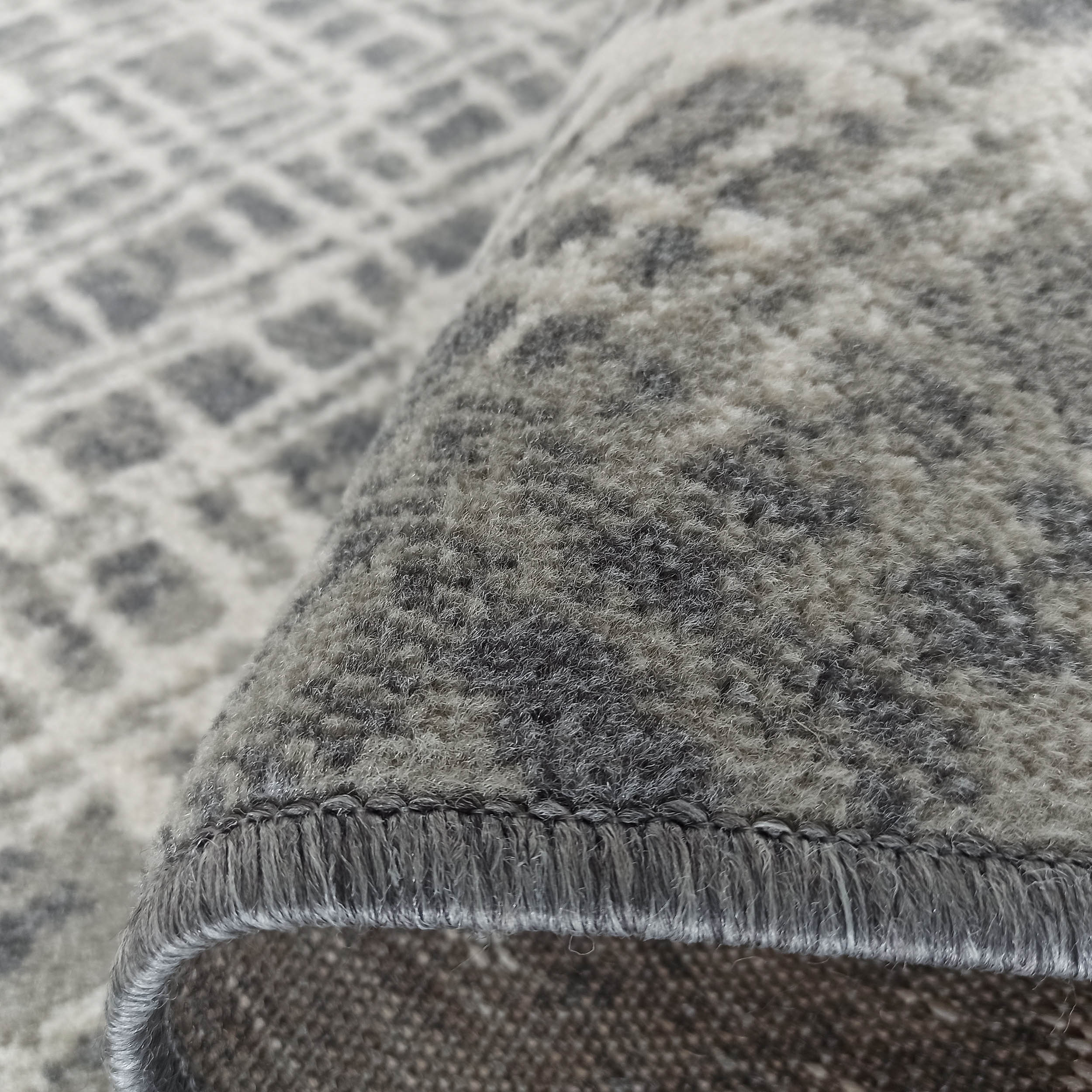 Gros plan sur un tapis texturé gris et blanc, partiellement roulé, révélant un motif abstrait et des mèches douces