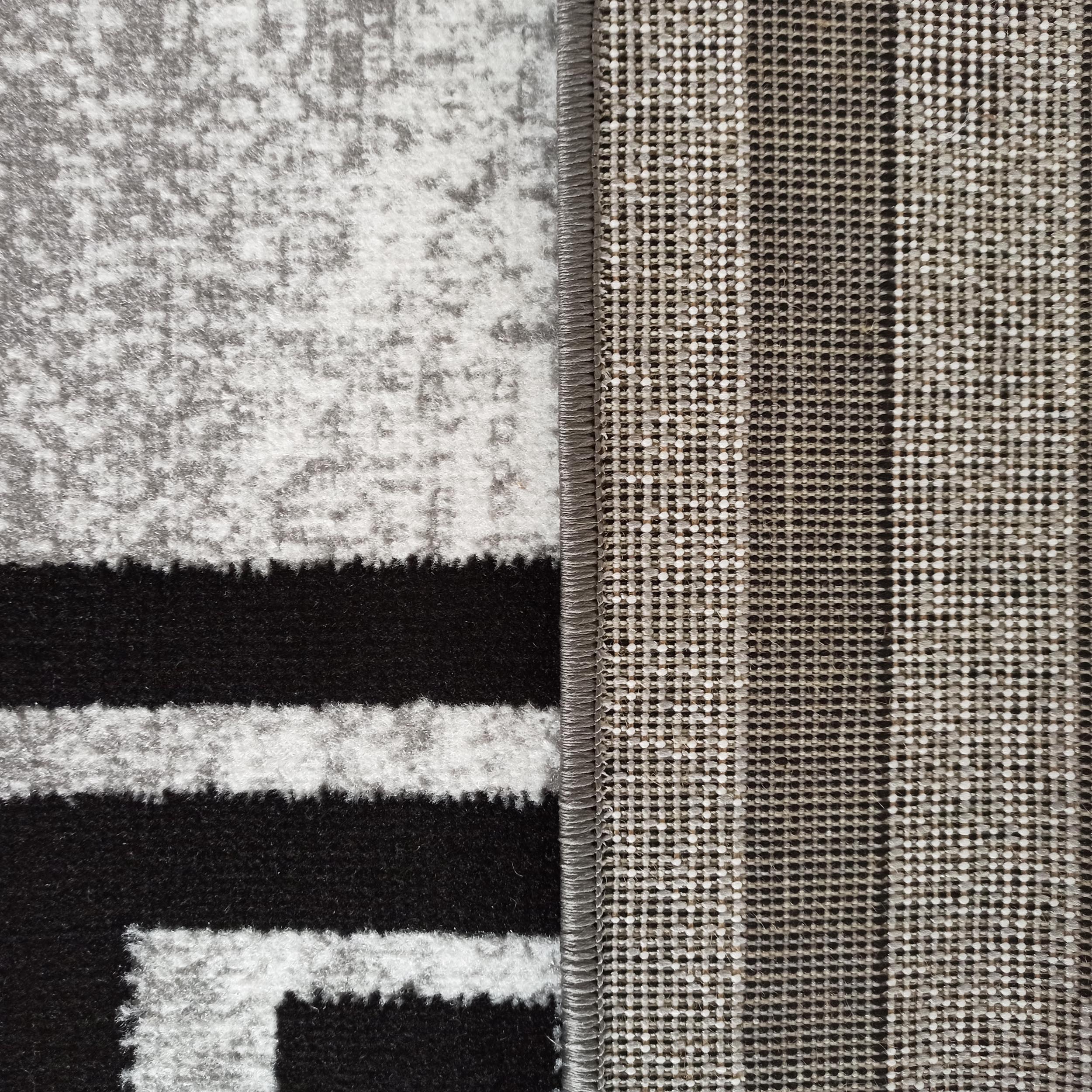 Gros plan sur un tapis, montrant la surface moelleuse à motifs noirs, blancs et gris chiné, le bord surjeté gris et l'envers tissé