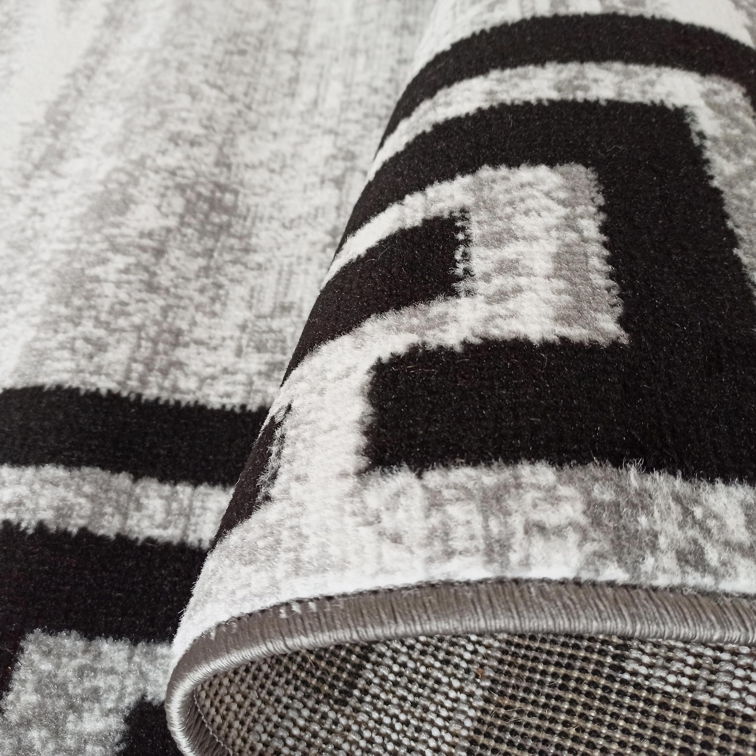Tapis moelleux enroulé, motifs géométriques noirs sur fond gris et blanc, bordure grise cousue visible