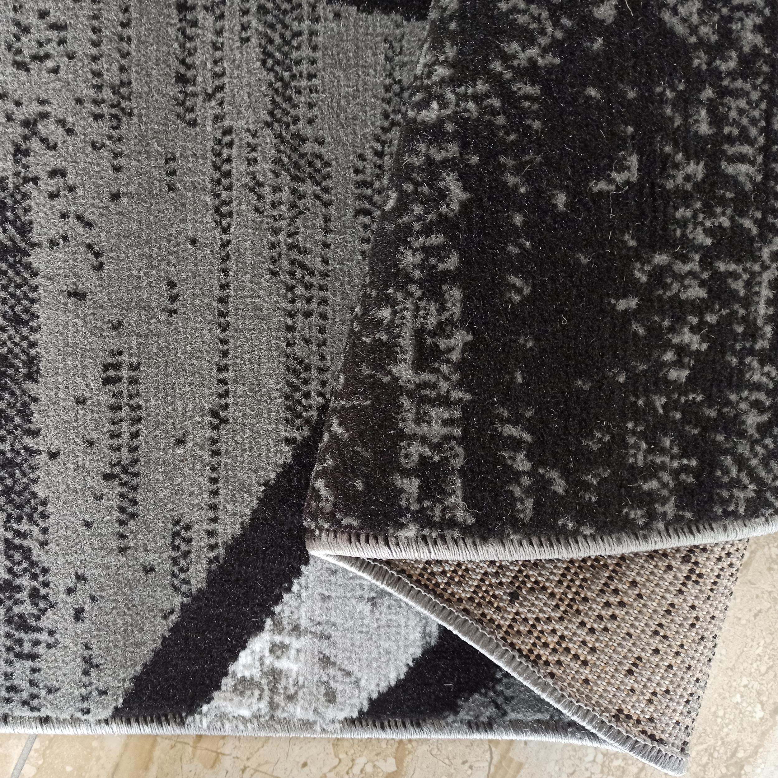 Tapis en textile à poils courts gris et noir avec un motif abstrait moucheté. Un coin est plié montrant le dessous tissé