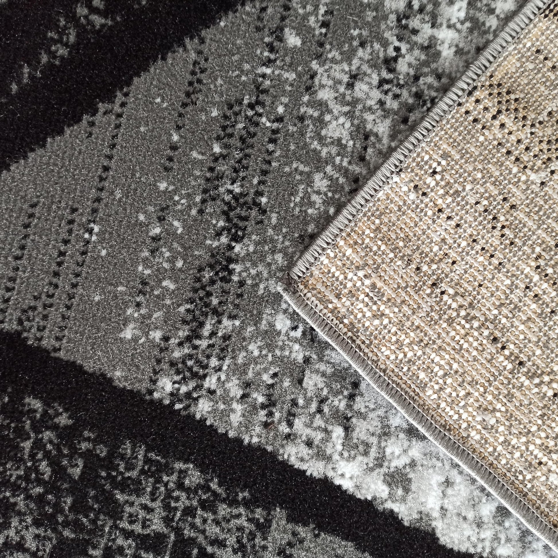 Tapis aux motifs abstraits mêlant noir, gris foncé et blanc. Son revers en tissu tissé beige et noir est visible sur un coin