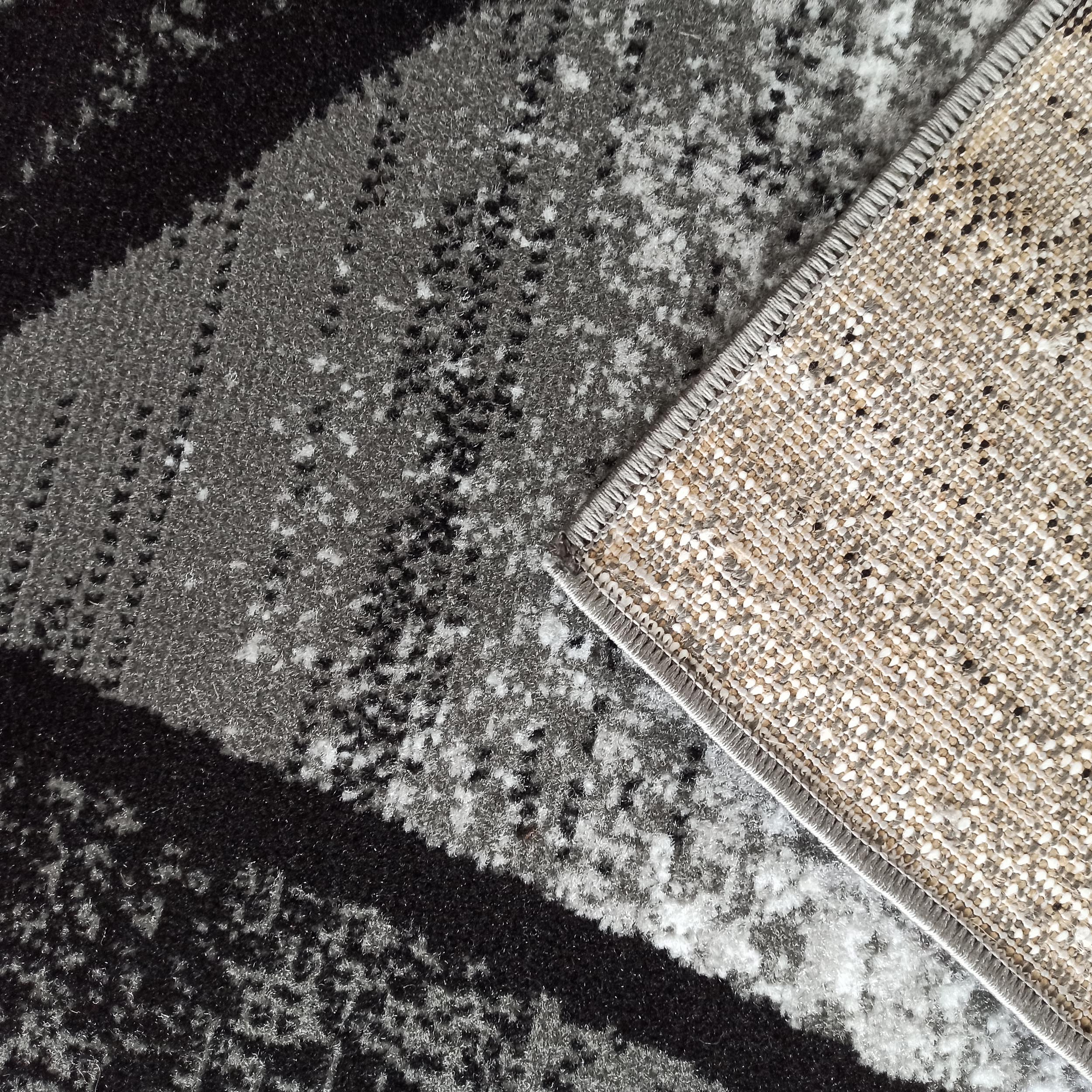 Tapis aux motifs abstraits mêlant noir, gris foncé et blanc. Son revers en tissu tissé beige et noir est visible sur un coin