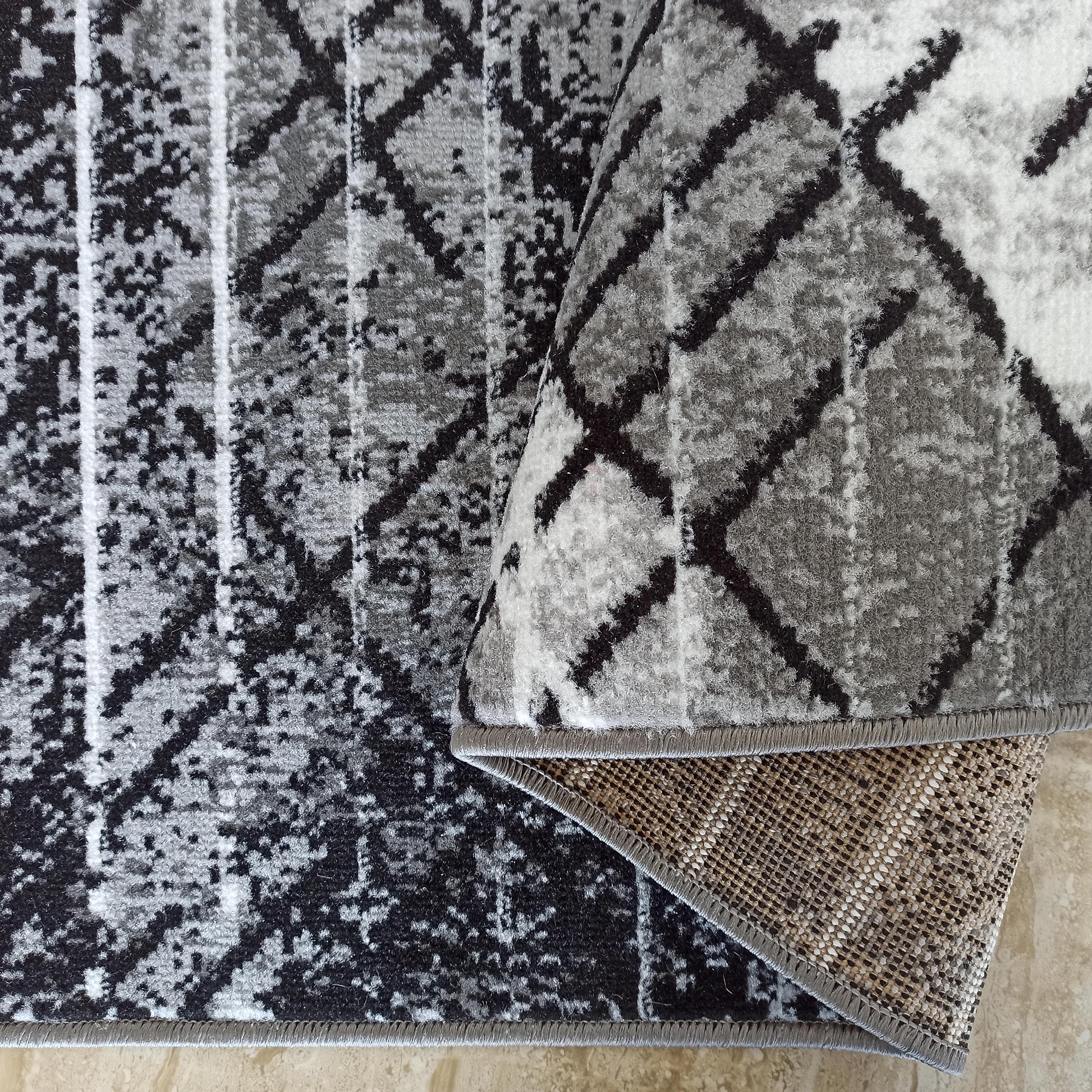 Tapis texturé aux motifs abstraits noirs, gris et blancs, partiellement roulé, montrant son envers tissé et son bord ourlé