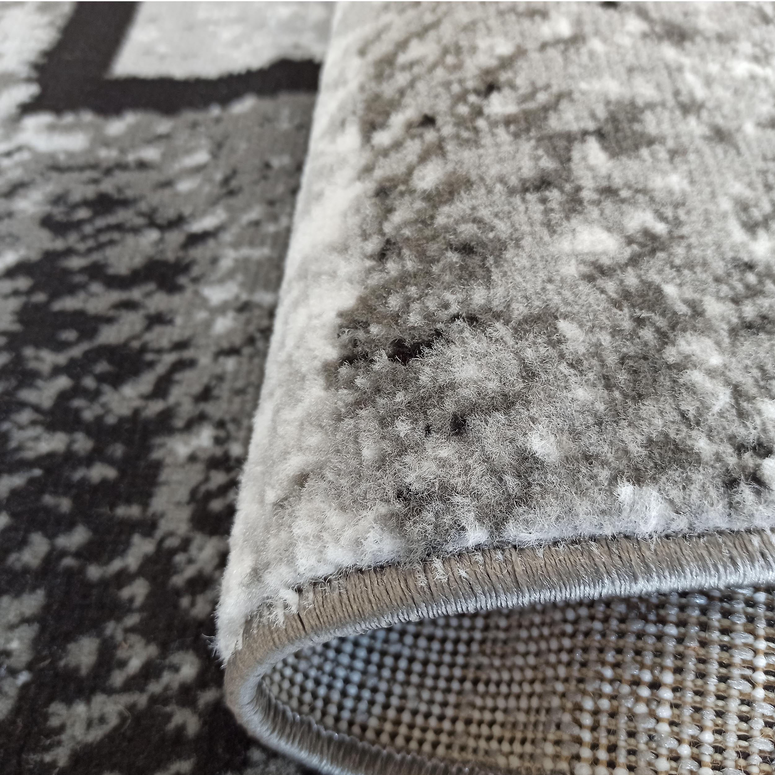 Tapis doux gris et blanc aux motifs abstraits, partiellement enroulé révélant son envers texturé quadrillé et ses bords finis