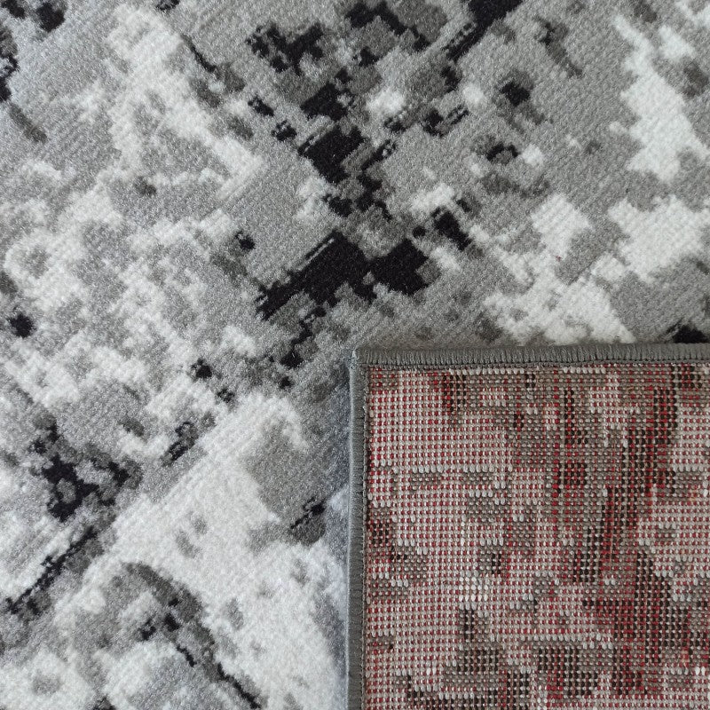 Tapis à motifs abstraits gris, noir et blanc, avec un coin relevé montrant le dos rouge et tissé