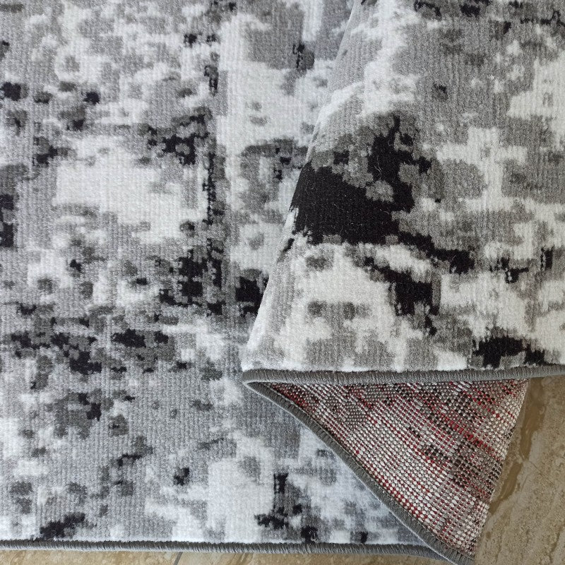 Tapis doux gris, blanc et noir aux motifs abstraits, avec un coin replié montrant son envers maillé rouge