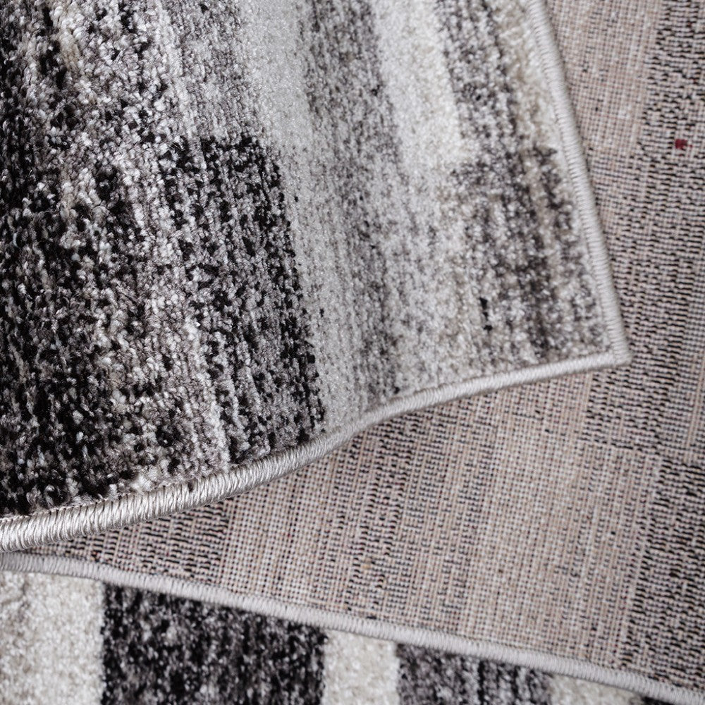 Tapis aux teintes de gris, noir et blanc cassé avec un motif abstrait, montrant son côté velours et son envers tissé beige