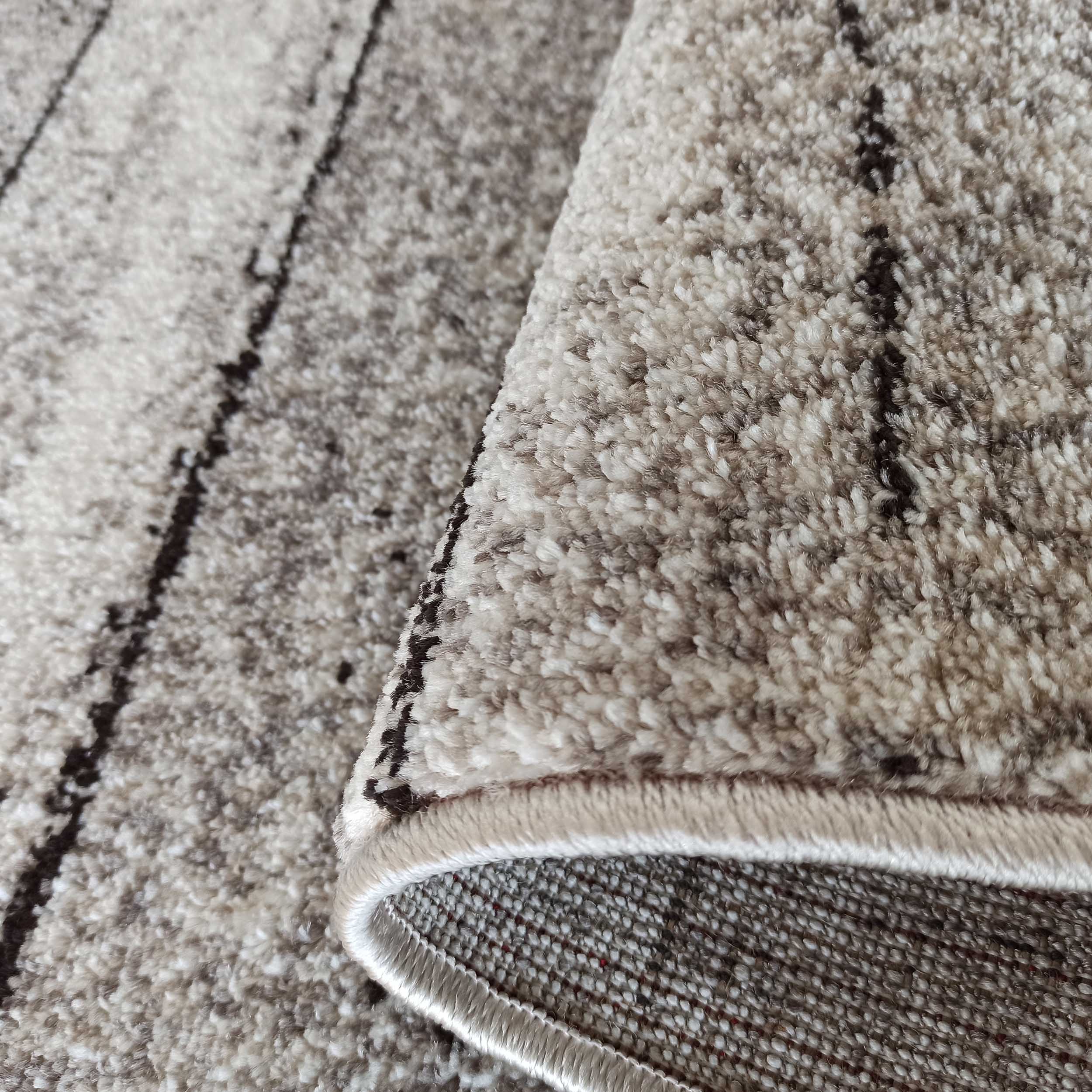 Tapis doux et texturé, principalement beige et crème, avec des motifs foncés irréguliers. Un bord est relevé, montrant la finition et l'envers tissé