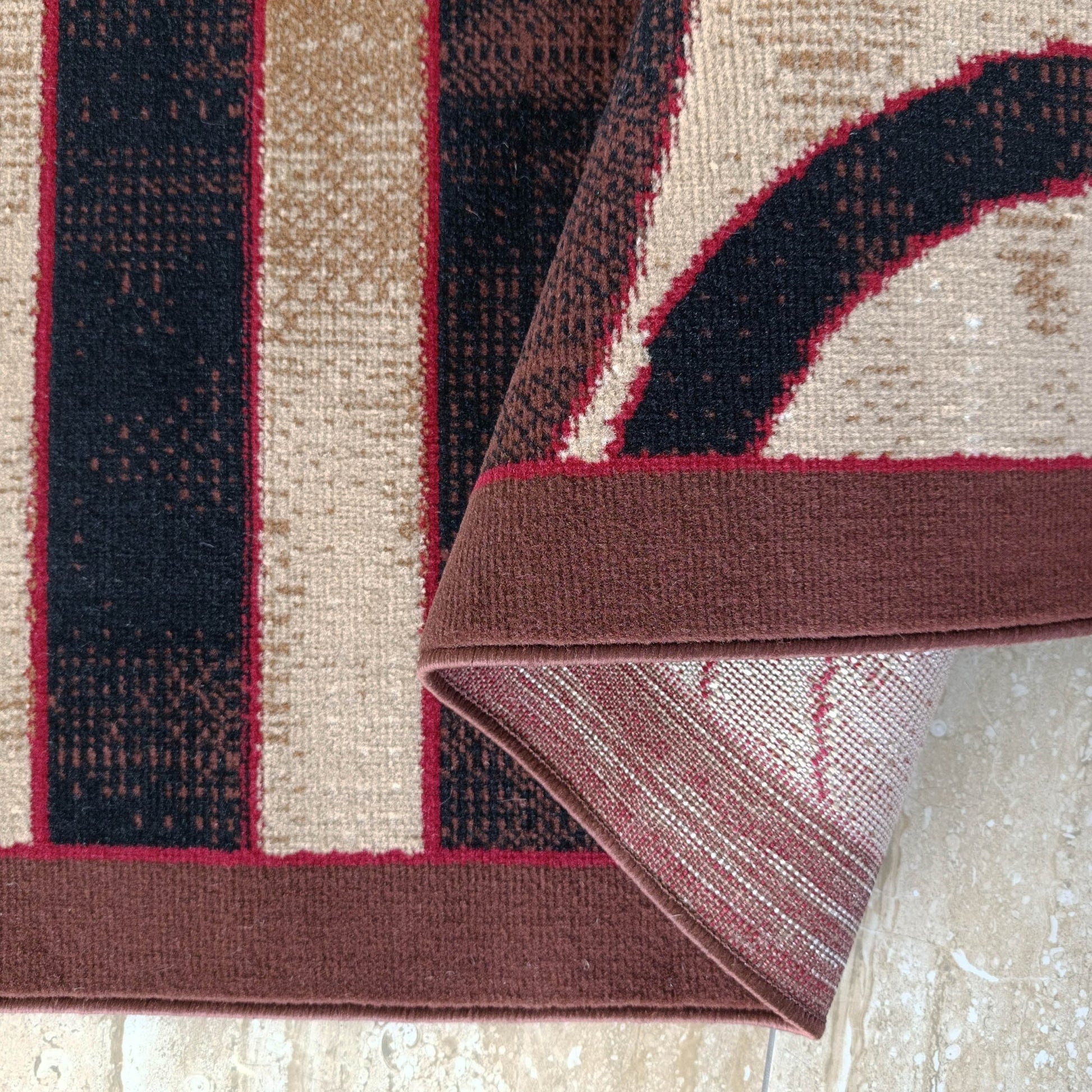 Motifs géométriques beiges, marron foncé et rouges sur un tapis replié, révélant son envers tissé et un sol clair