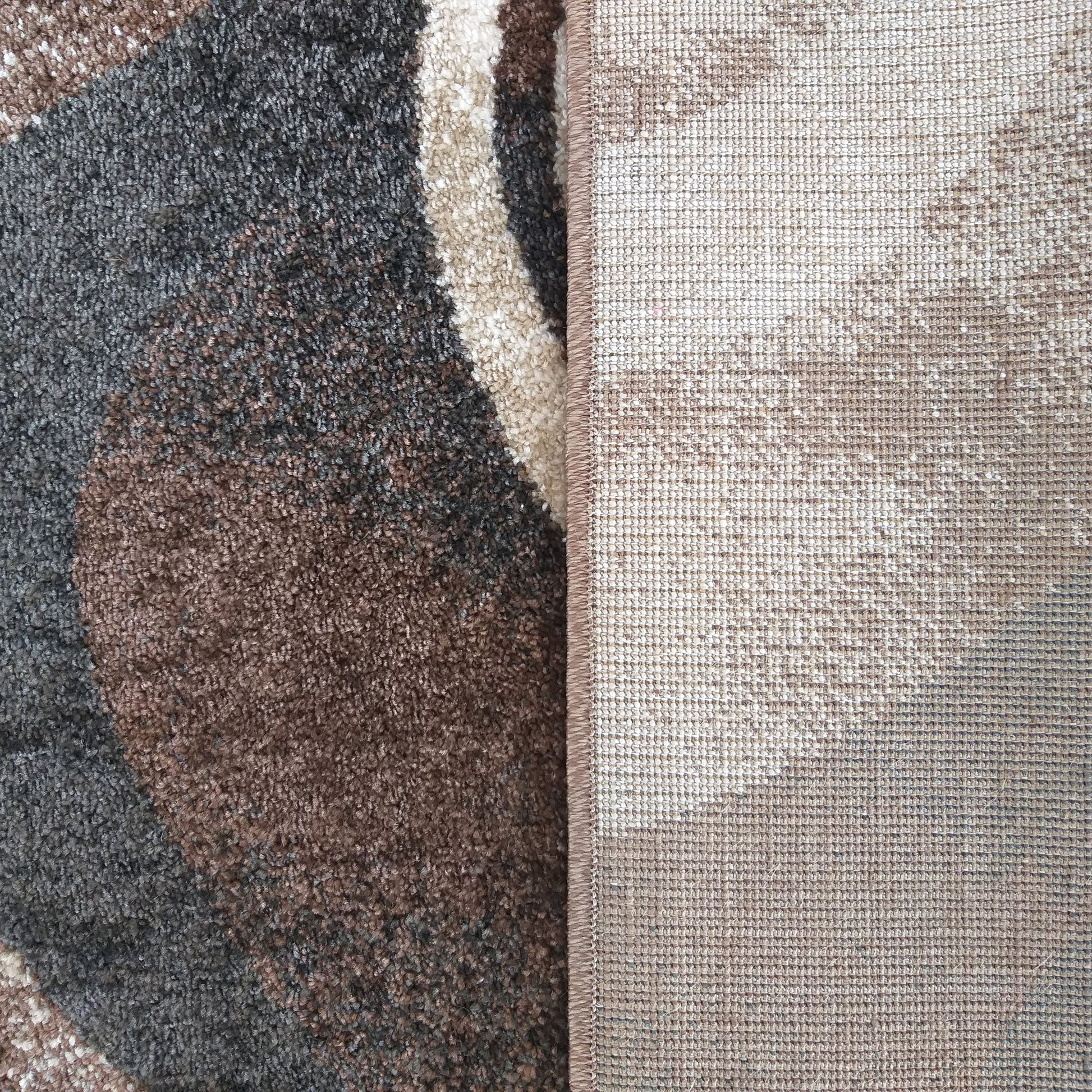 Tapis à la texture dense et aux motifs abstraits marron, gris, blanc cassé. L'envers beige tissé du tapis et sa bordure cousue sont visibles