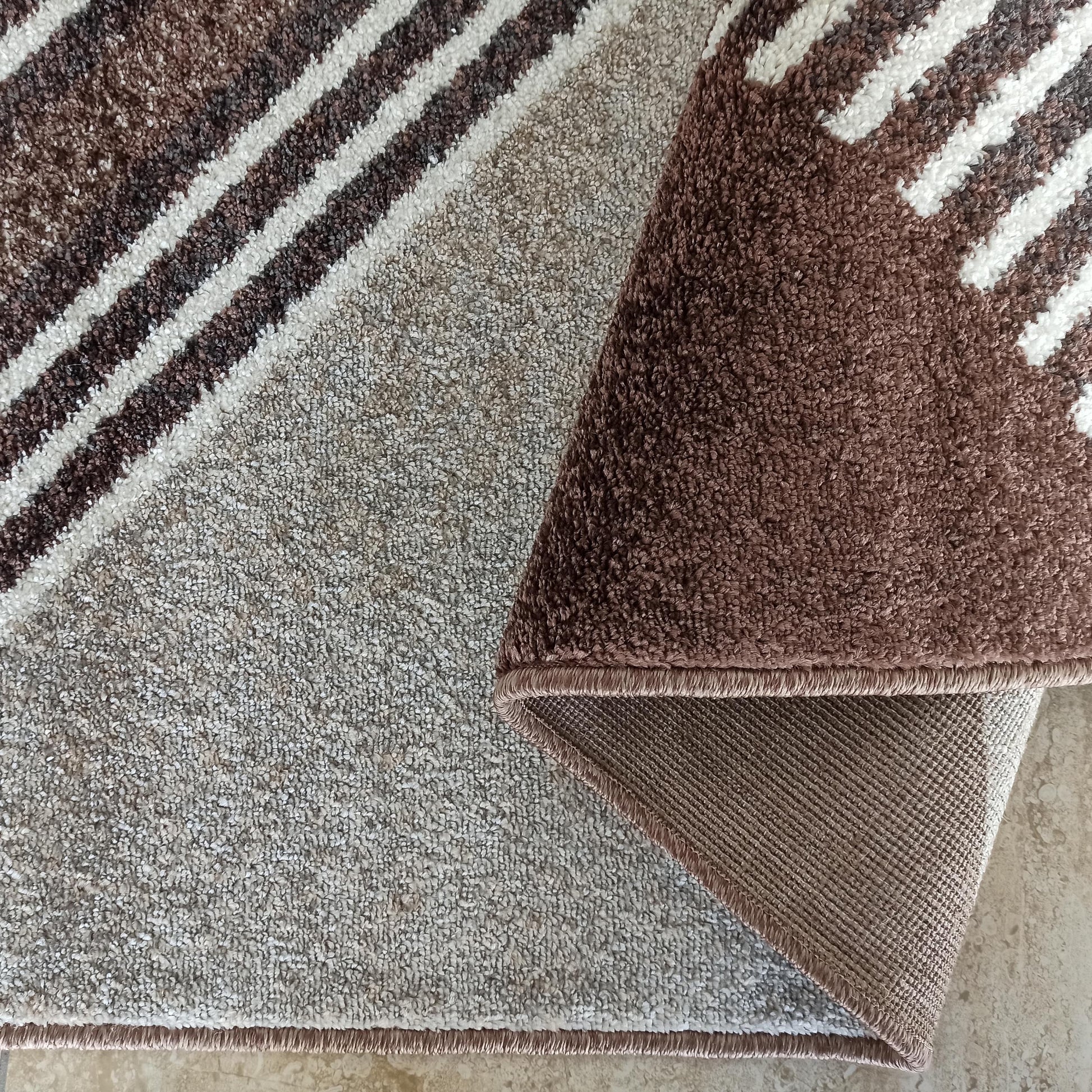Tapis moelleux aux motifs de rayures diagonales marron foncé, blanches et beiges. Une partie est roulée, révélant son envers tissé et sa bordure