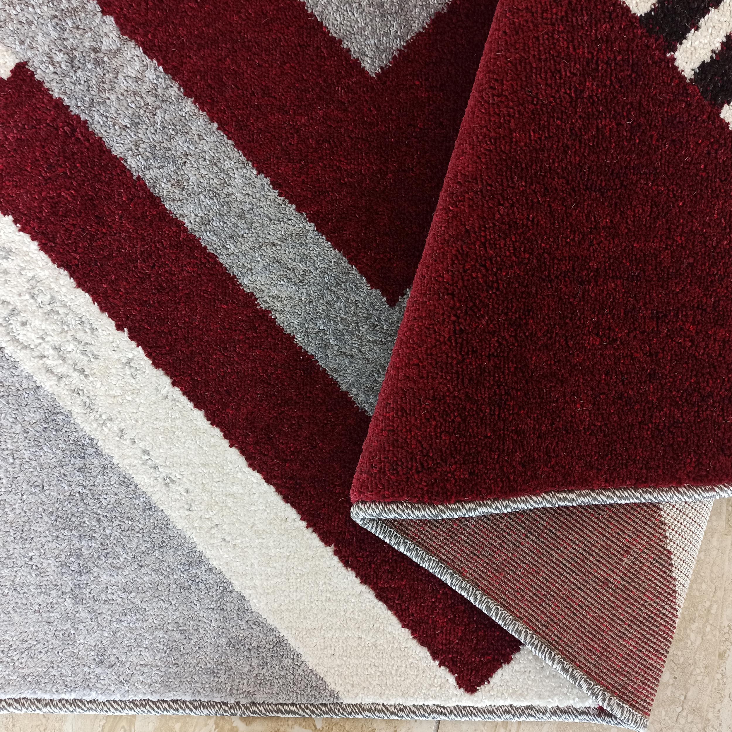 Tapis en textile moelleux à motifs géométriques : bandes obliques rouge bordeaux, grises et blanches, replié