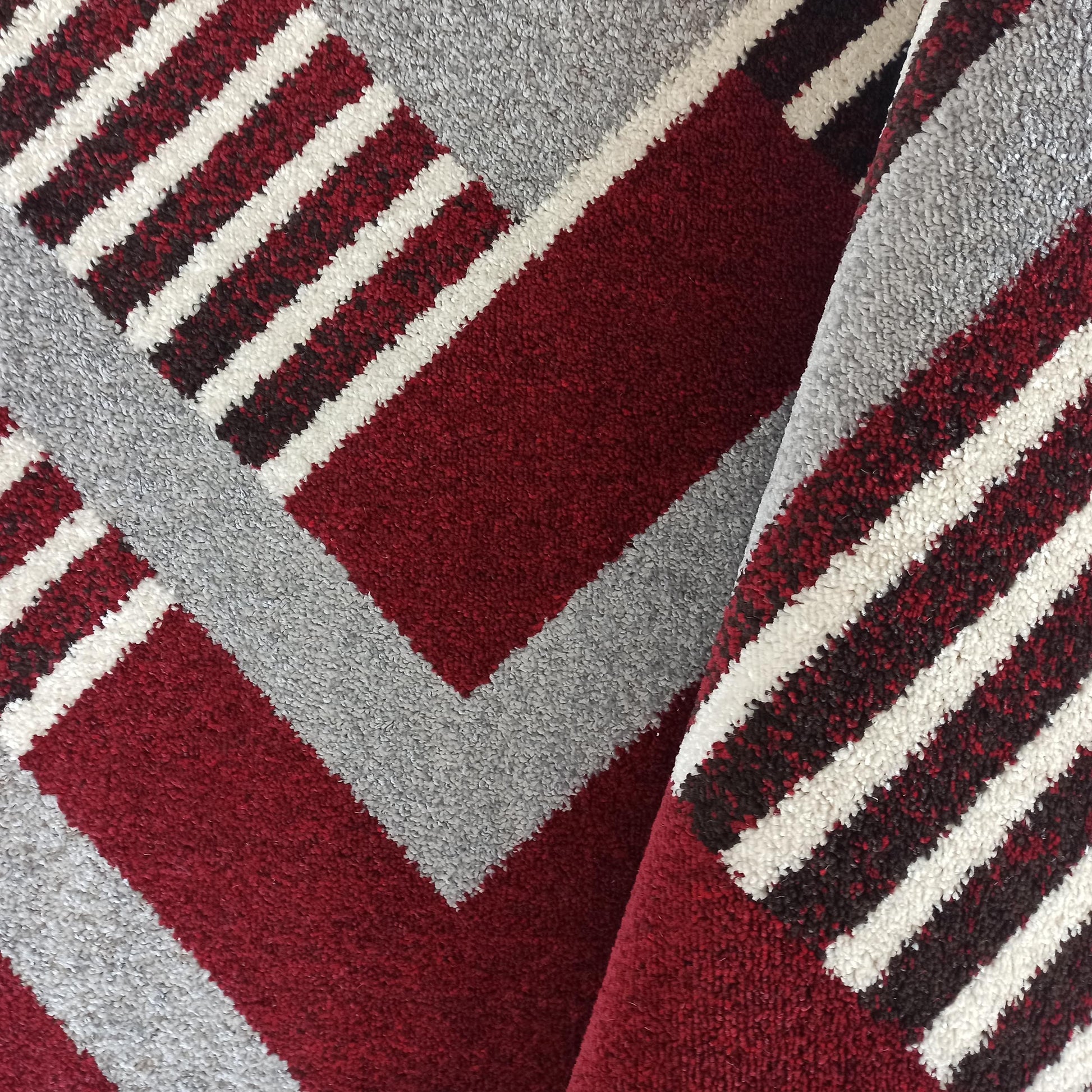 Tapis aux motifs géométriques et aux couleurs rouge bordeaux, gris clair et blanc, avec une texture moelleuse visible sur le pli