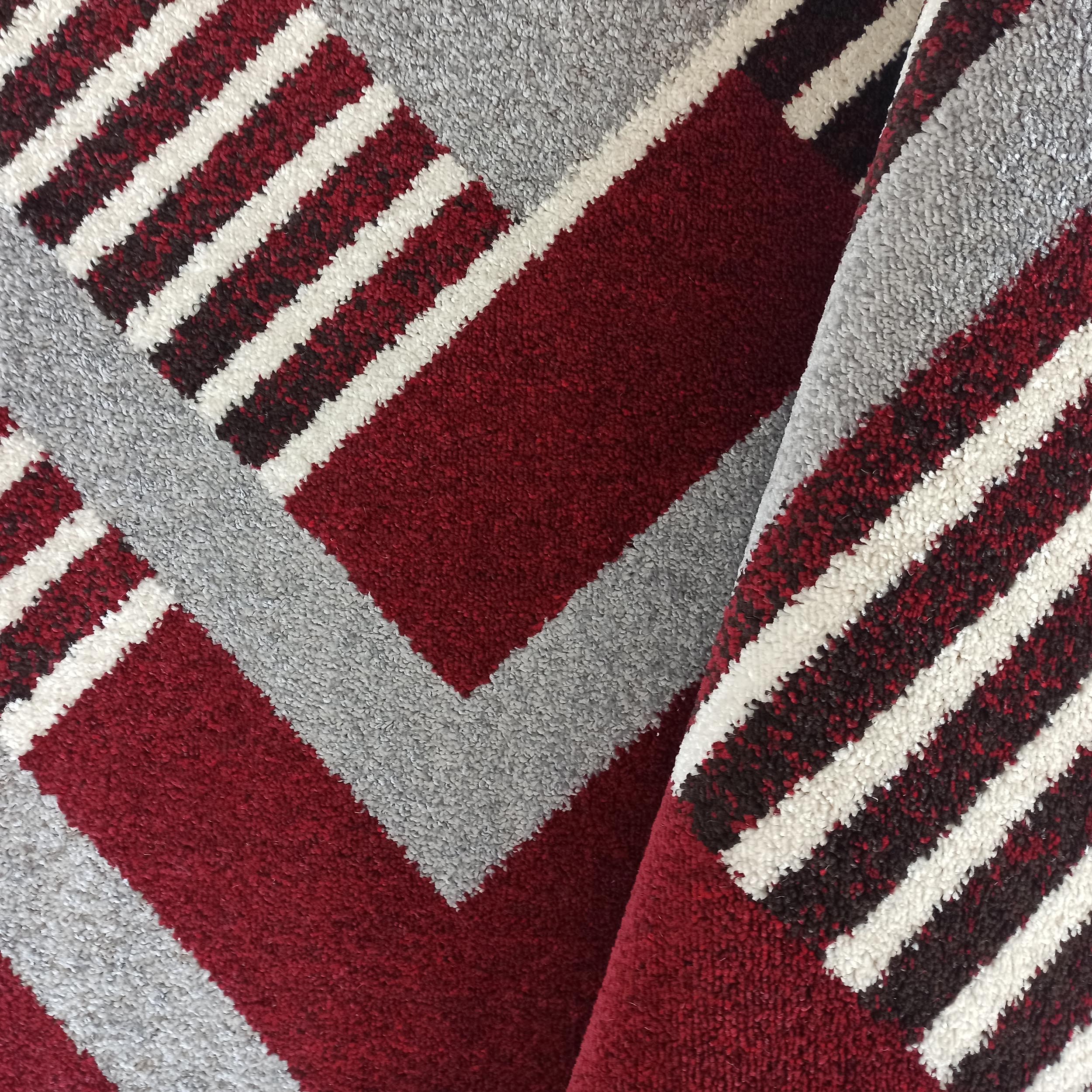 Tapis aux motifs géométriques et aux couleurs rouge bordeaux, gris clair et blanc, avec une texture moelleuse visible sur le pli