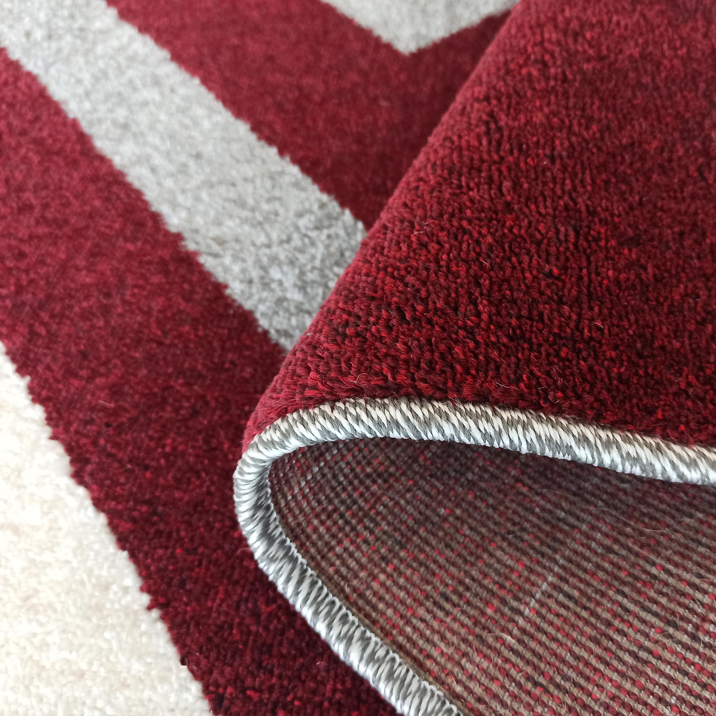 Tapis doux aux motifs géométriques bordeaux et blanc. Une partie relevée dévoile sa bordure surjetée grise et son envers tissé