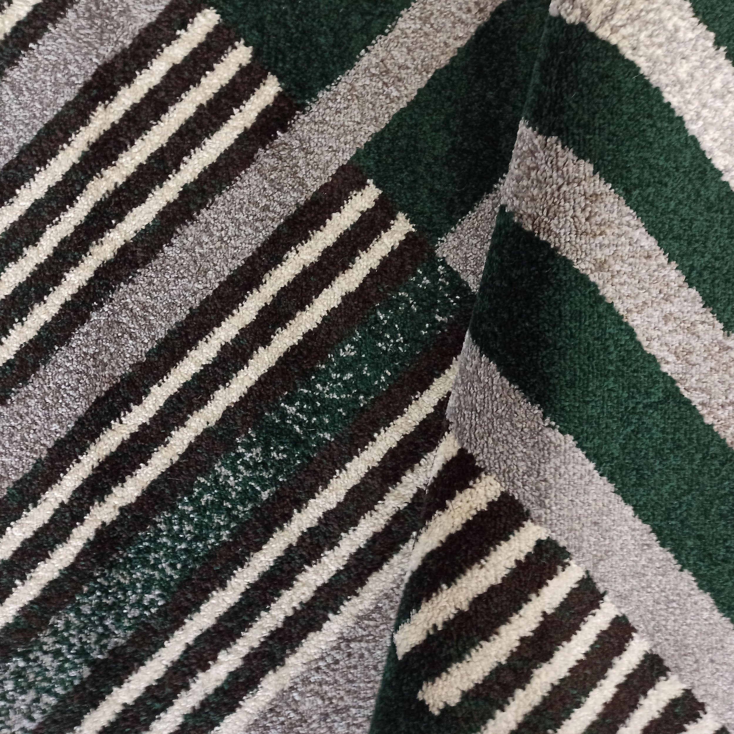 Tapis à poils longs avec un motif de bandes diagonales épaisses et fines en vert foncé, gris clair, marron et crème, avec un pli visible