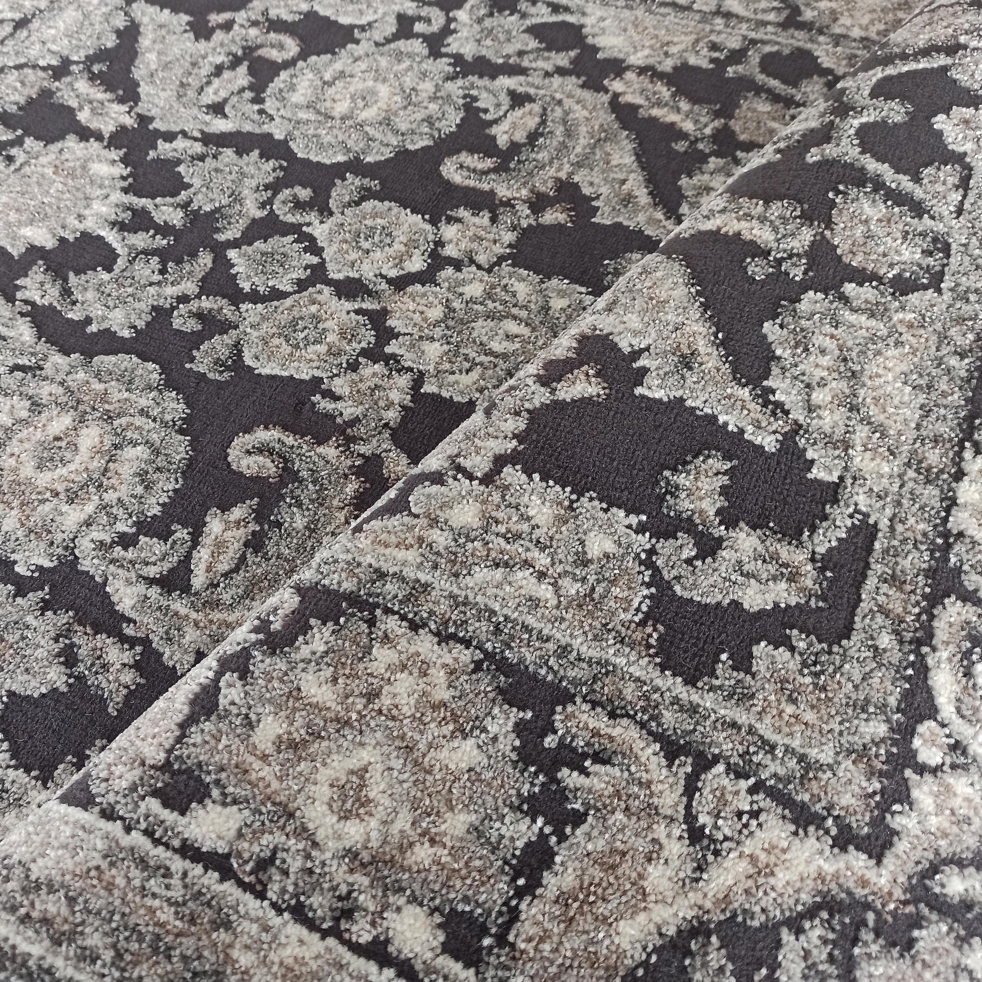 Tapis en textile bouclé gris foncé avec des motifs ornementaux clairs et beiges au relief moelleux, légèrement plié
