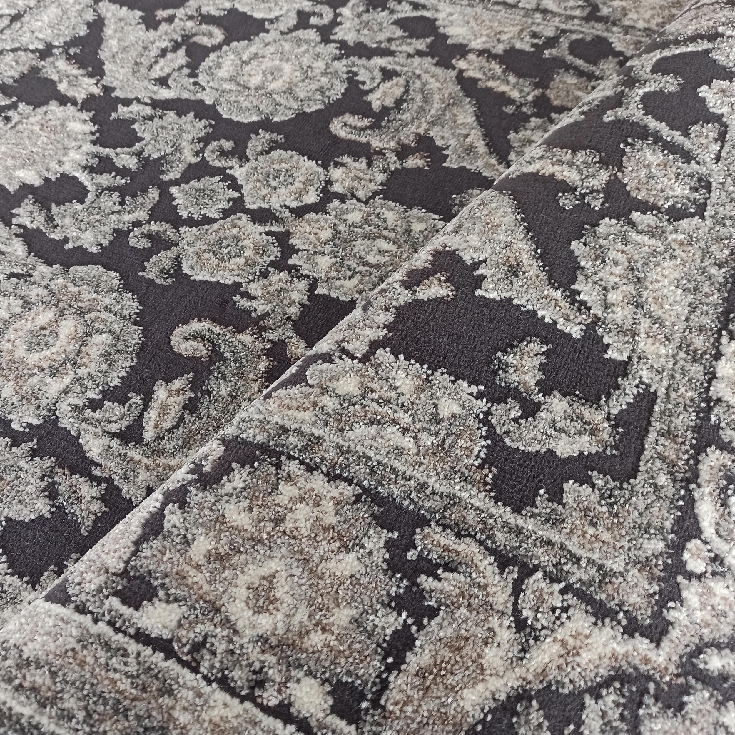 Tapis en textile bouclé gris foncé avec des motifs ornementaux clairs et beiges au relief moelleux, légèrement plié