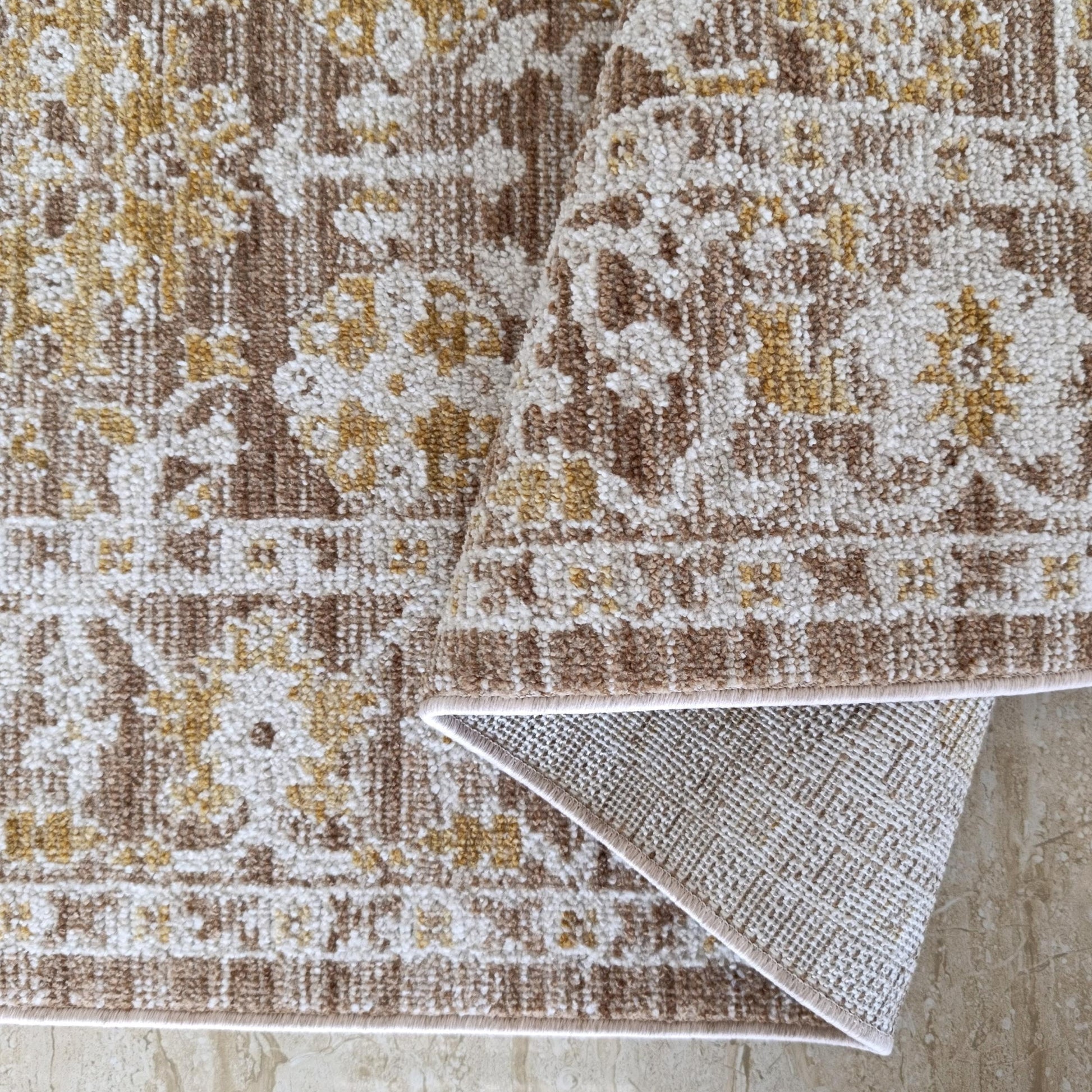 Tapis texturé crème, marron clair, or à motifs ornementaux, roulé sur carrelage beige marbré, envers tissé apparent