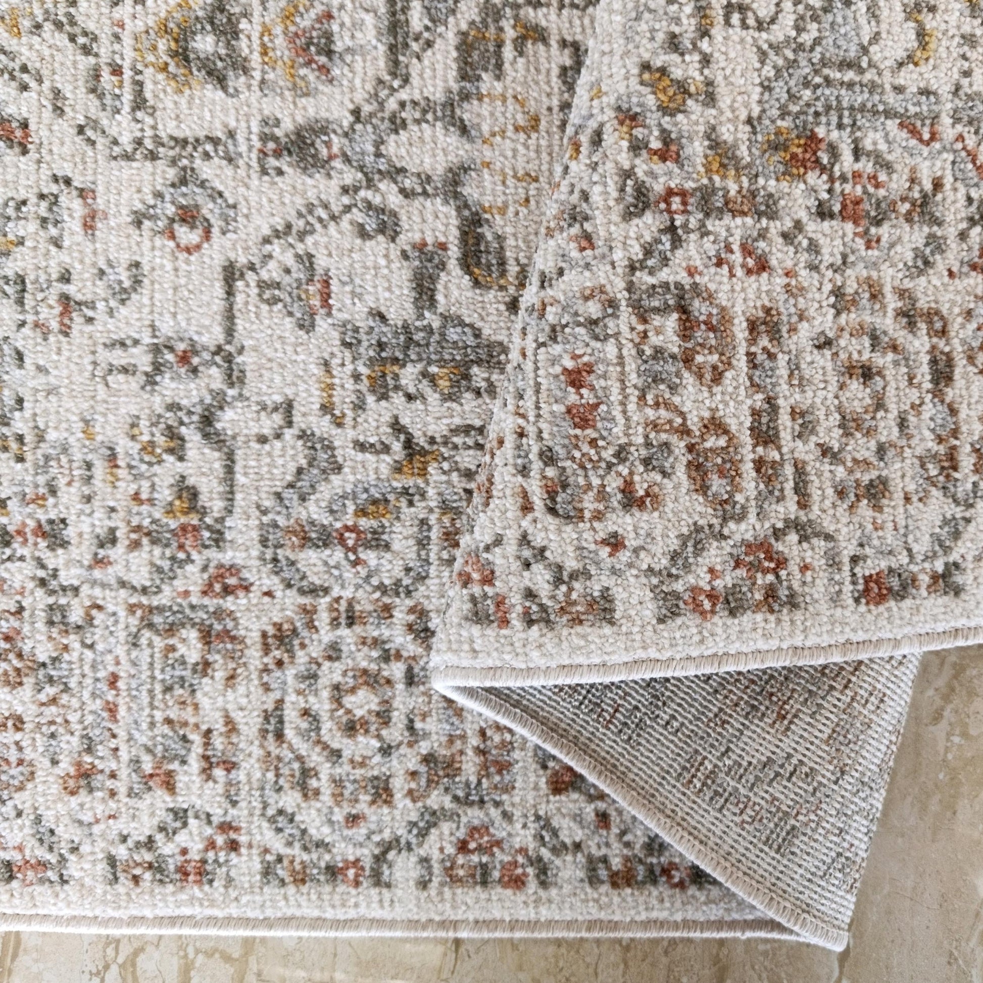 Tapis moelleux crème, motifs gris, rouille et ocre, un coin replié montrant le tissage sur carrelage clair