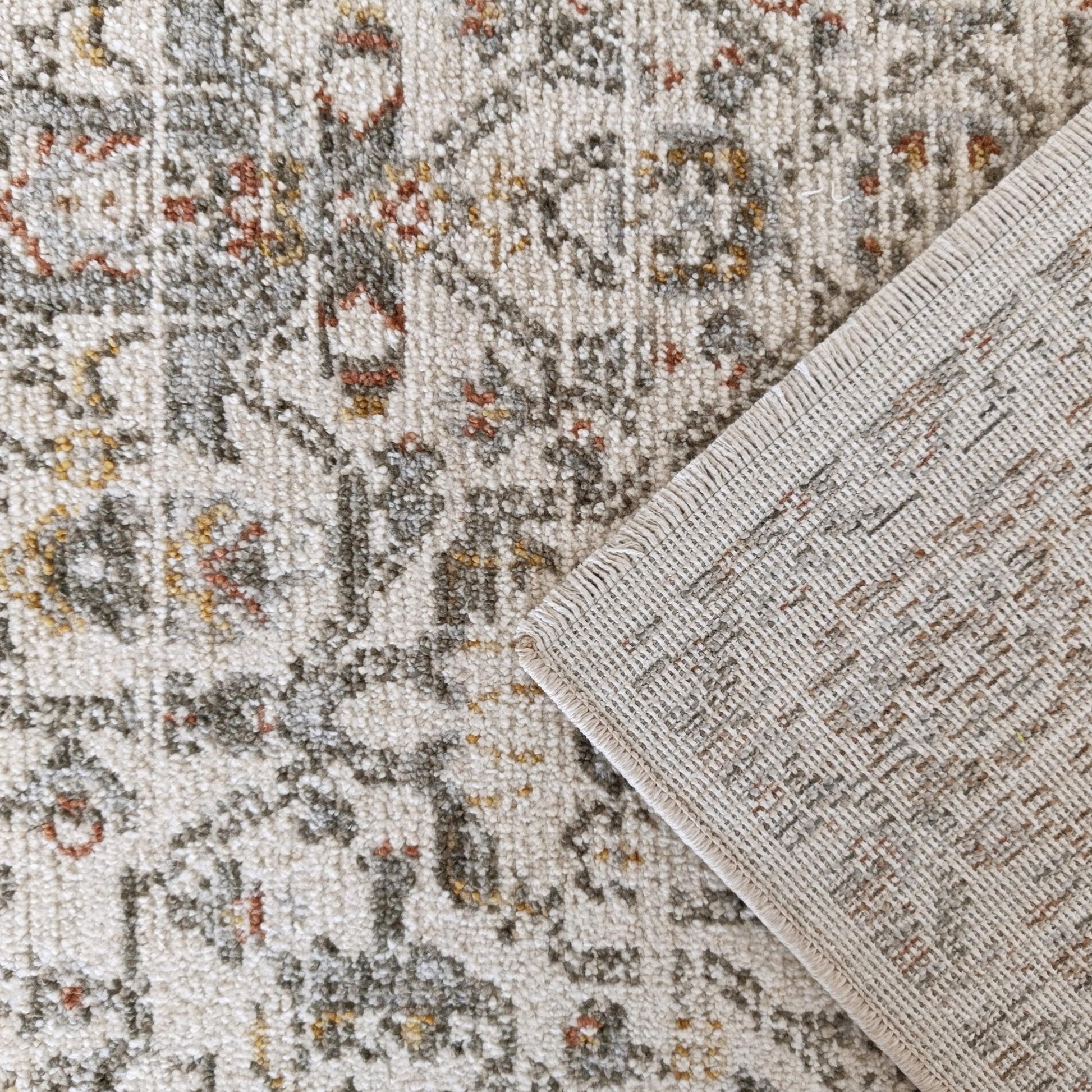 Un tapis crème à motifs ornementaux gris, rouille et jaune, son coin replié révélant un envers tissé beige et texturé
