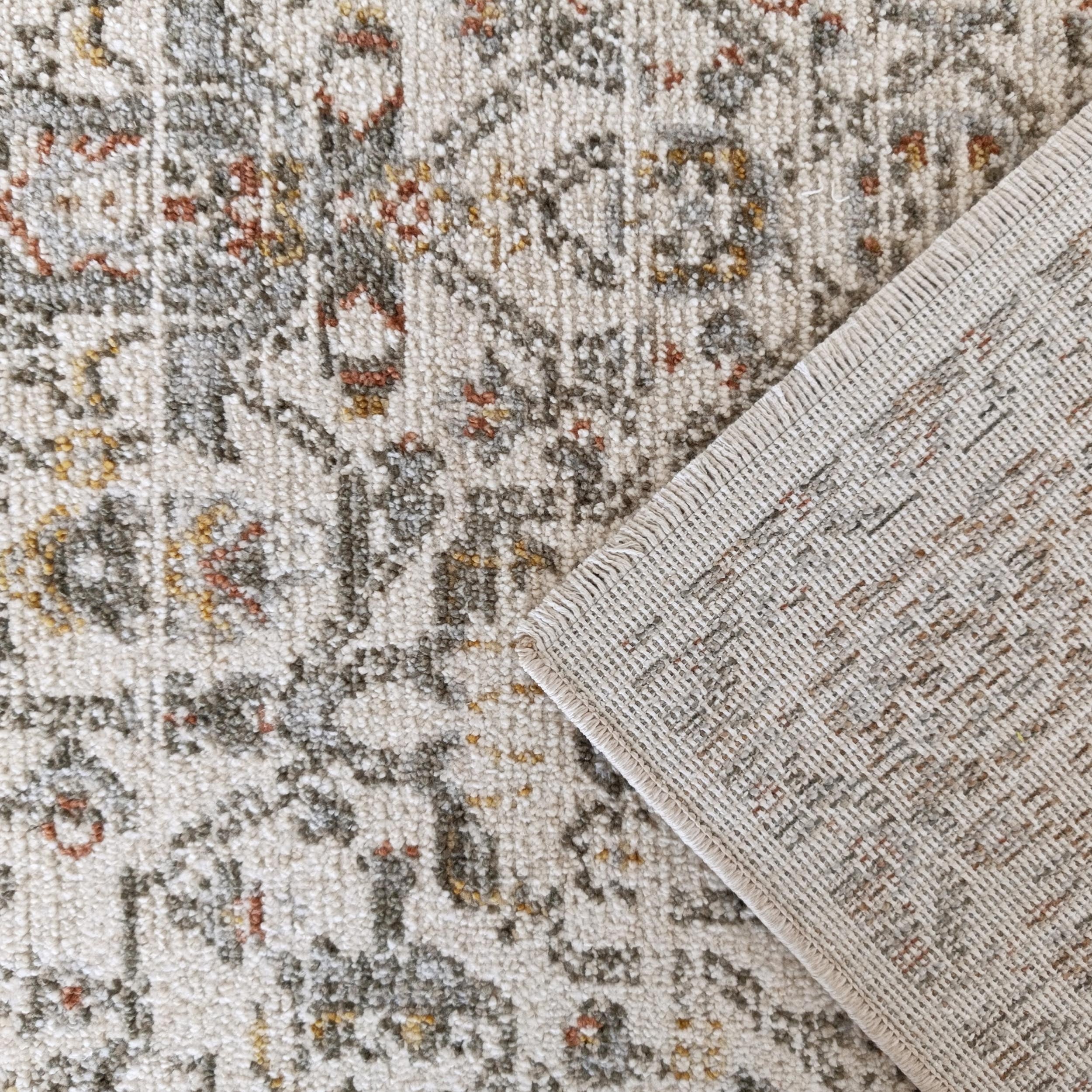 Un tapis crème à motifs ornementaux gris, rouille et jaune, son coin replié révélant un envers tissé beige et texturé