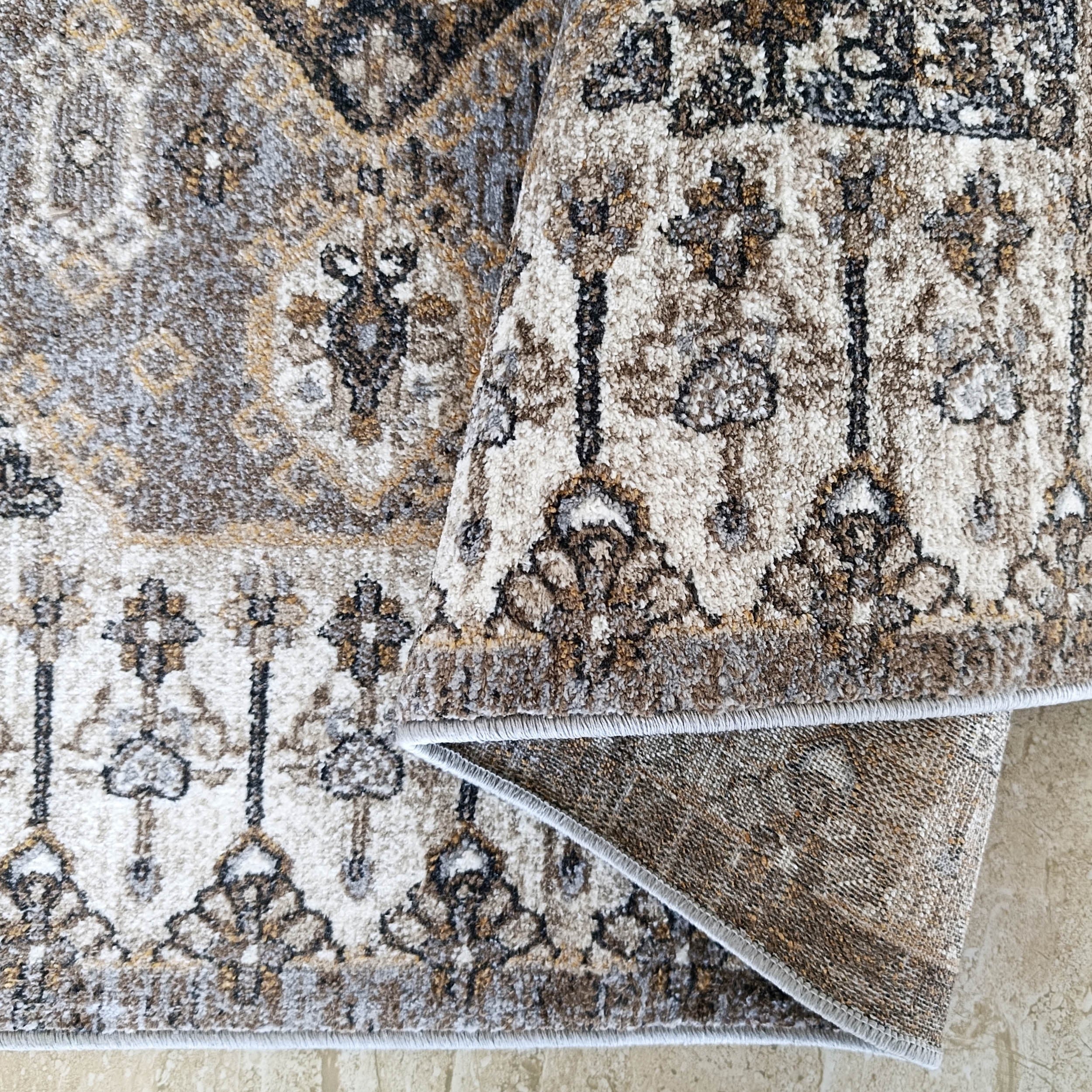 Tapis moelleux aux motifs ornementaux crème, gris, marron et doré, partiellement roulé sur un sol clair
