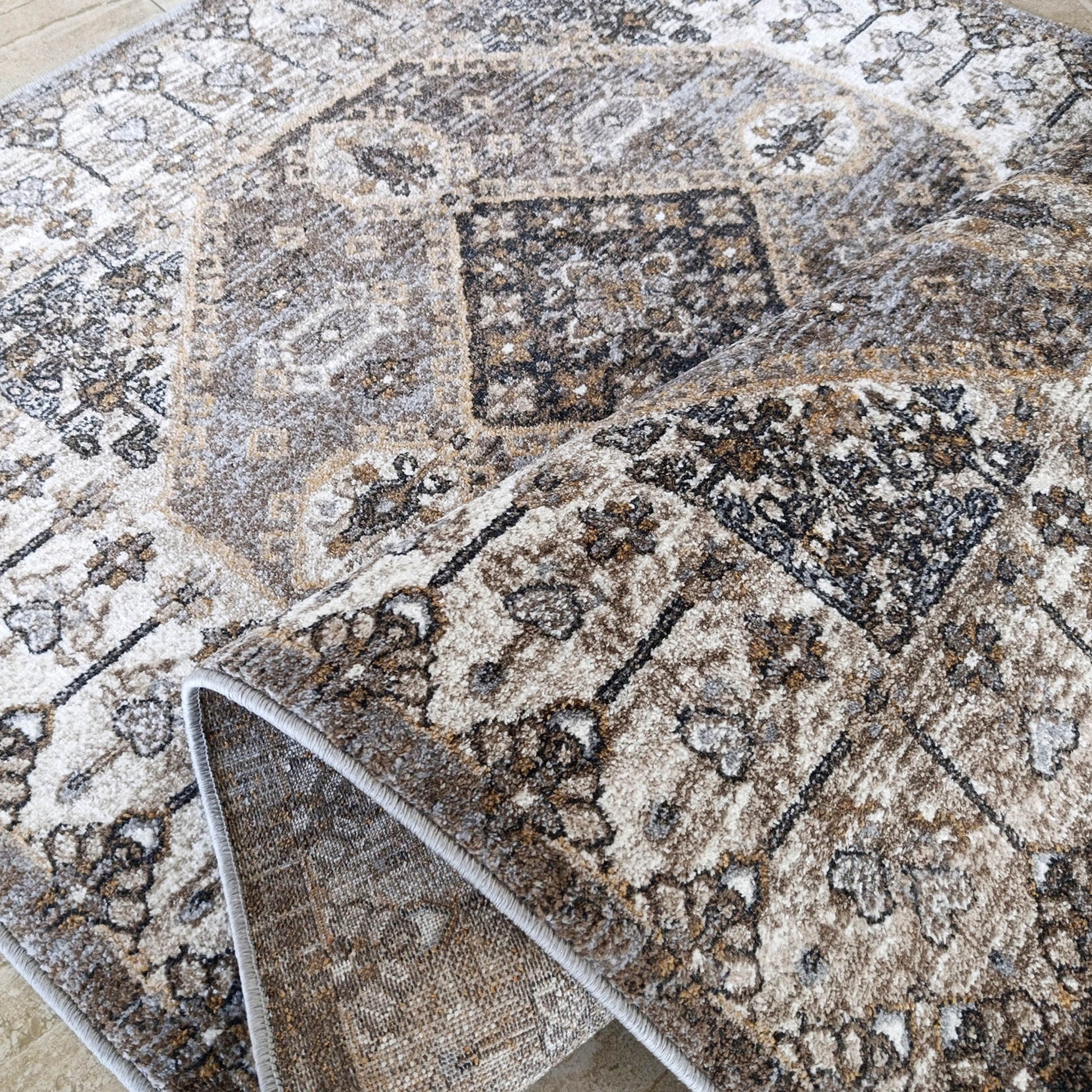 Tapis moelleux aux motifs ornementaux beige, marron, gris et noir, partiellement roulé sur un sol carrelé clair