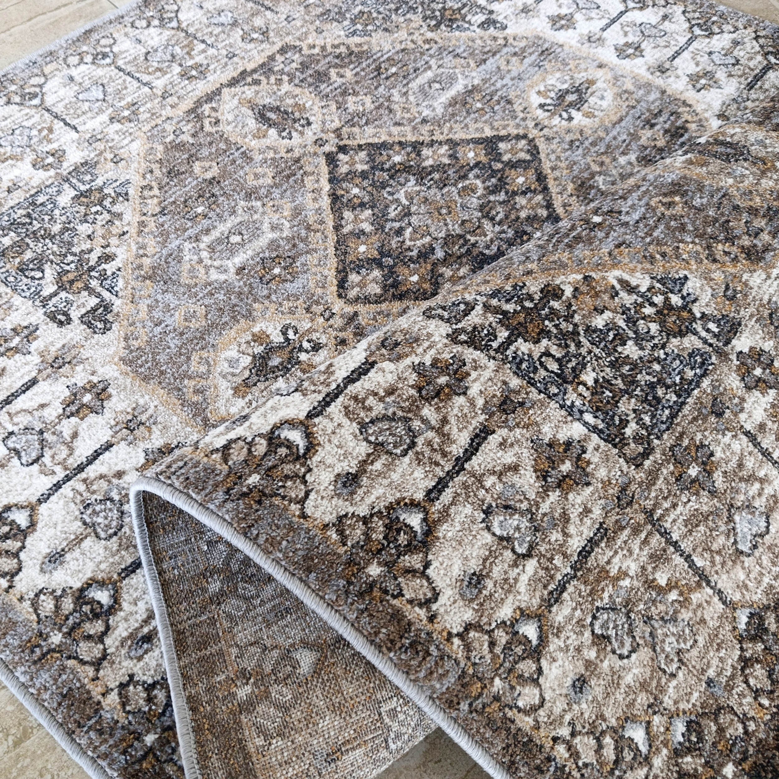 Tapis moelleux aux motifs ornementaux beige, marron, gris et noir, partiellement roulé sur un sol carrelé clair