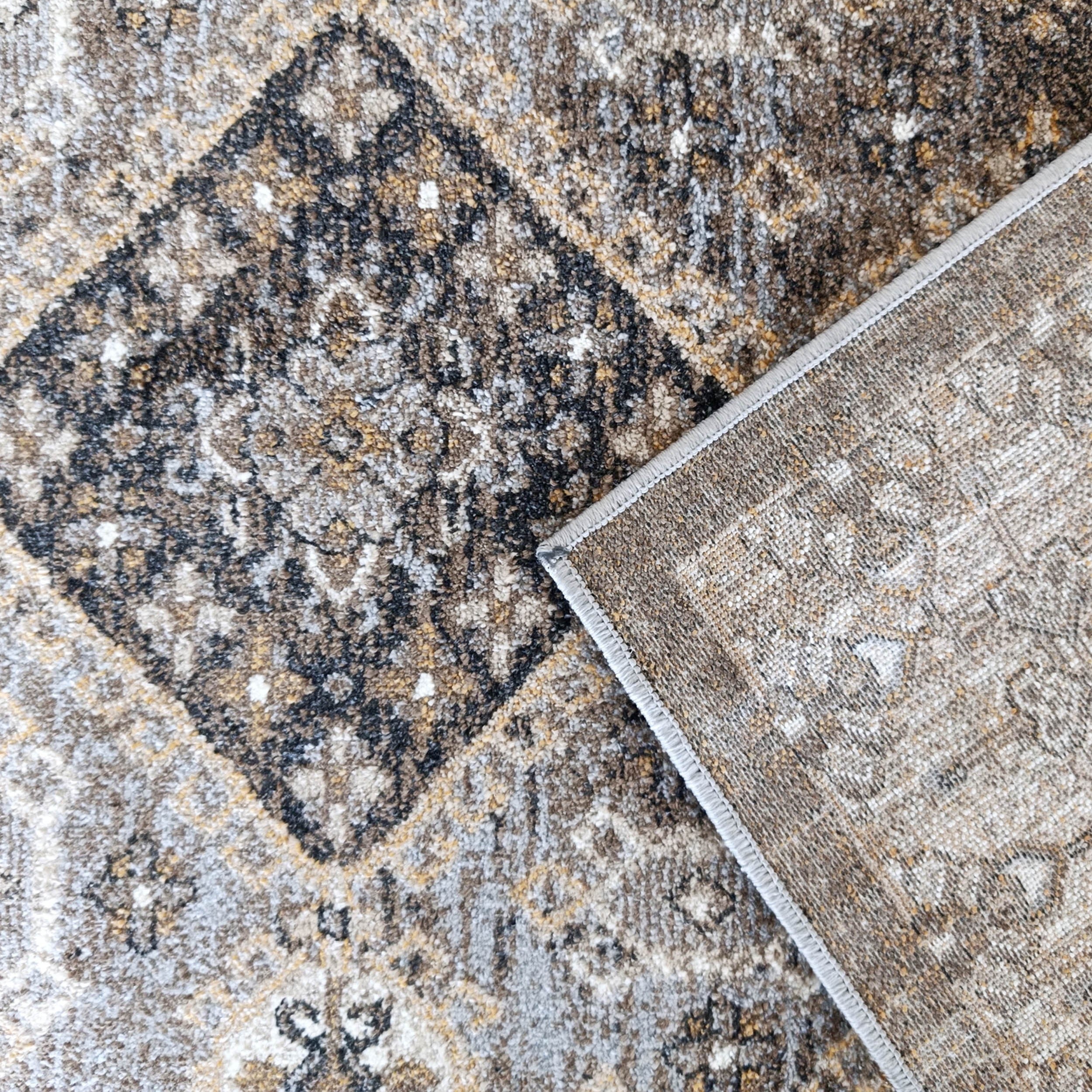 Tapis aux motifs géométriques marron, gris et doré, avec un coin replié montrant son envers tissé et son bord surpiqué