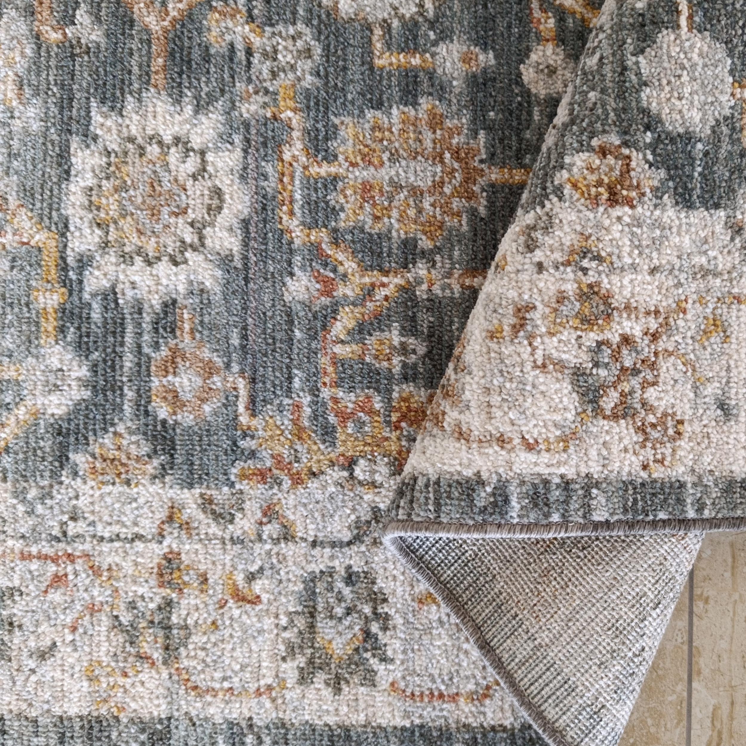 Tapis moelleux à motifs floraux ornementaux bleu-vert, crème, ocre et rouille, avec un coin relevé sur un sol carrelé
