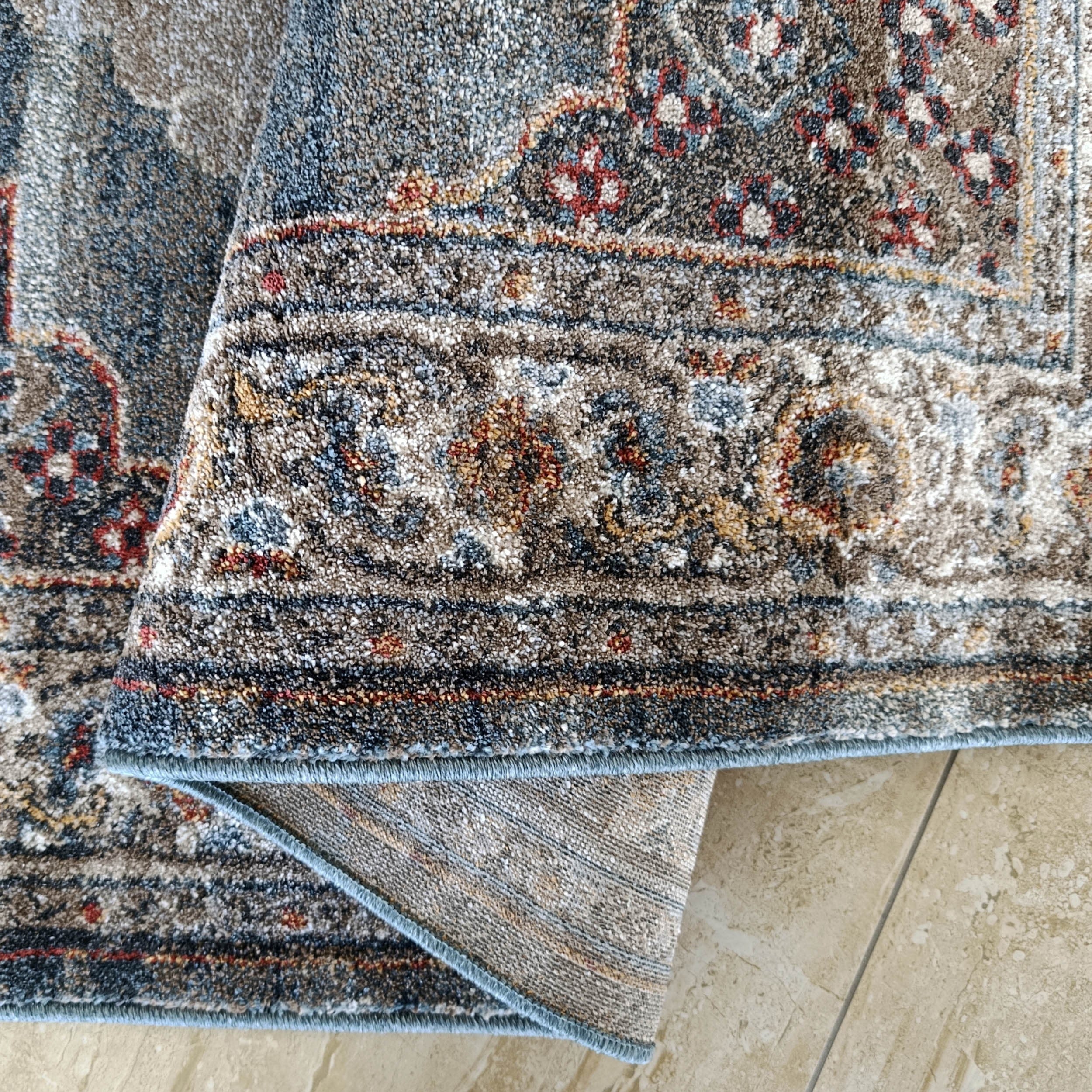 Tapis moelleux à motifs traditionnels gris, bleus, beiges, rouges, à bord bleu clair, plié sur carrelage beige
