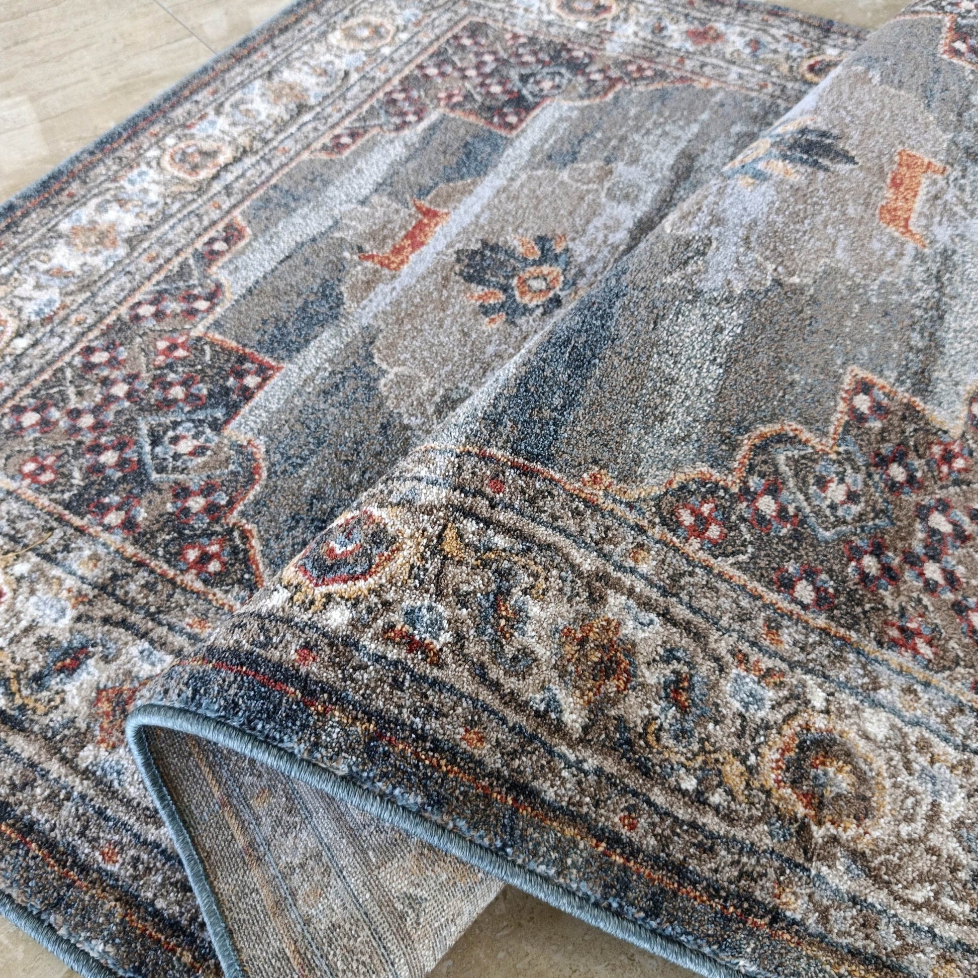 Tapis doux gris, bleu et ocre avec motifs traditionnels floraux et géométriques, partiellement enroulé sur un carrelage beige